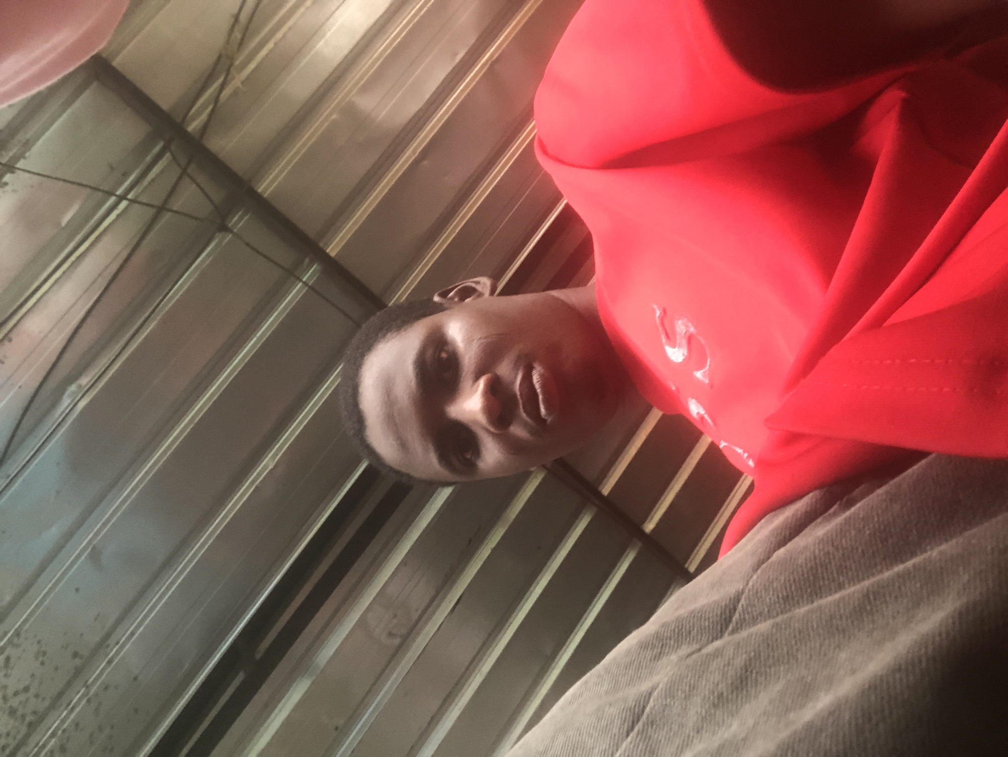 A low-angle, close-up shot features a Black man with short, dark hair wearing a bright red shirt. The camera is positioned below the man, looking up at his face. The background consists of corrugated metal siding. There is nothing to indicate this image was taken in Ado, Nigeria. There are no discernable objects, interactions, or actions.