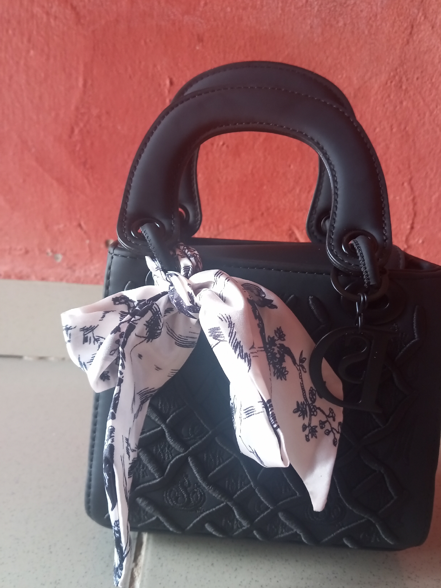 This is a medium shot, eye-level photograph of a black, quilted handbag. The bag has a structured silhouette with a top handle and a metal charm with the letters "CD" attached to the side. A small, white scarf with a black floral pattern is tied to the bag's handle. The bag is resting on a light-colored surface, possibly a countertop or table. Behind the bag, a textured, reddish-orange wall is visible. The lighting is soft and appears to be natural. The image seems to be taken indoors, with no discernible clues about the time of day or weather. There are no people visible in the image, and the focus is entirely on the handbag. The overall impression is one of an item of fashion presented in a simple, uncluttered setting.