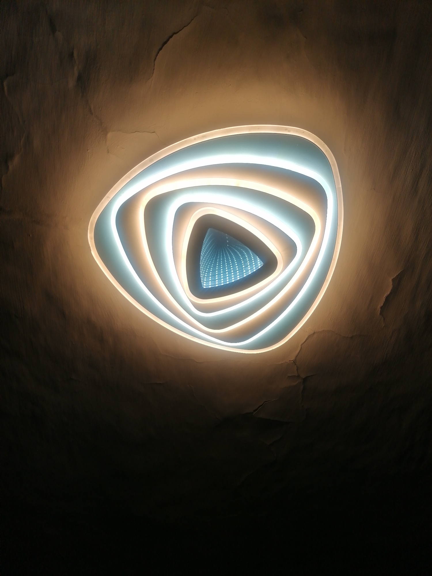 A modern, triangular-shaped ceiling light fixture with an infinity mirror effect is centrally located on a textured, cracked ceiling. The fixture emits a warm white and cool blue light, creating concentric rings that recede into the mirror, giving the illusion of depth. The ceiling itself is a dark, earthy brown with visible cracks and imperfections, suggesting an older or weathered building. The overall lighting is dim, with the fixture being the primary light source. The image does not contain any people or specific environmental details that would indicate its location within Serebriansk, Kazakhstan.