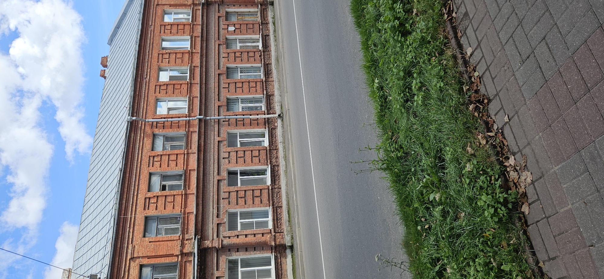 This image depicts a two-story brick building with a metal roof, located along a street in Vitebsk, Belarus. The building features numerous windows on both floors, some with white frames and curtains, and others with visible interior spaces. The brickwork of the building shows decorative horizontal and vertical brick detailing. A downspout runs vertically along the front of the building. In front of the building is a paved road with a white dividing line. A strip of green grass and some fallen leaves separates the road from a paved sidewalk constructed with red and gray interlocking bricks. The sky above is blue with scattered white clouds. No people are visible in the scene.