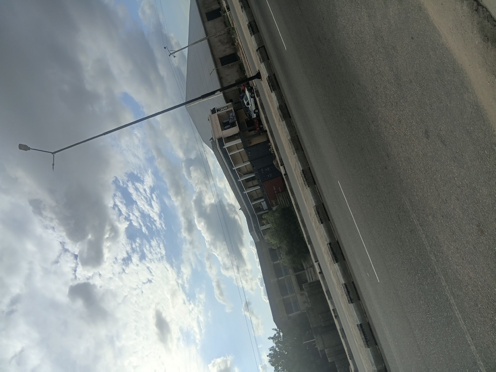 The image depicts a roadside view in Jalingo, Nigeria, under a partially cloudy sky. The foreground features a two-lane asphalt road with a single white lane marker. A concrete curb system, marked with dark rectangular patterns, separates the road from the adjacent shoulder.
A prominent black street light pole with a single primary light fixture and a shorter secondary arm extends upwards from the right side of the road. Further right, a wooden utility pole with strung power lines is visible. Beyond these, a large building with a broad, light-colored pitched roof and dark walls, consistent with an industrial or warehouse structure, occupies the background.
To the left of the large structure, a multi-story building with a light-colored facade, balconies, and a sloped roof is present. A white pickup truck and a partially visible red pickup truck are parked on the shoulder in front of this building, adjacent to multiple dark shipping containers. Further left, another multi-story building with balconies and a weathered exterior is partially obscured by dense green foliage. The sky above displays a mix of white and grey clouds with intermittent patches of blue. No people are visible in the scene.