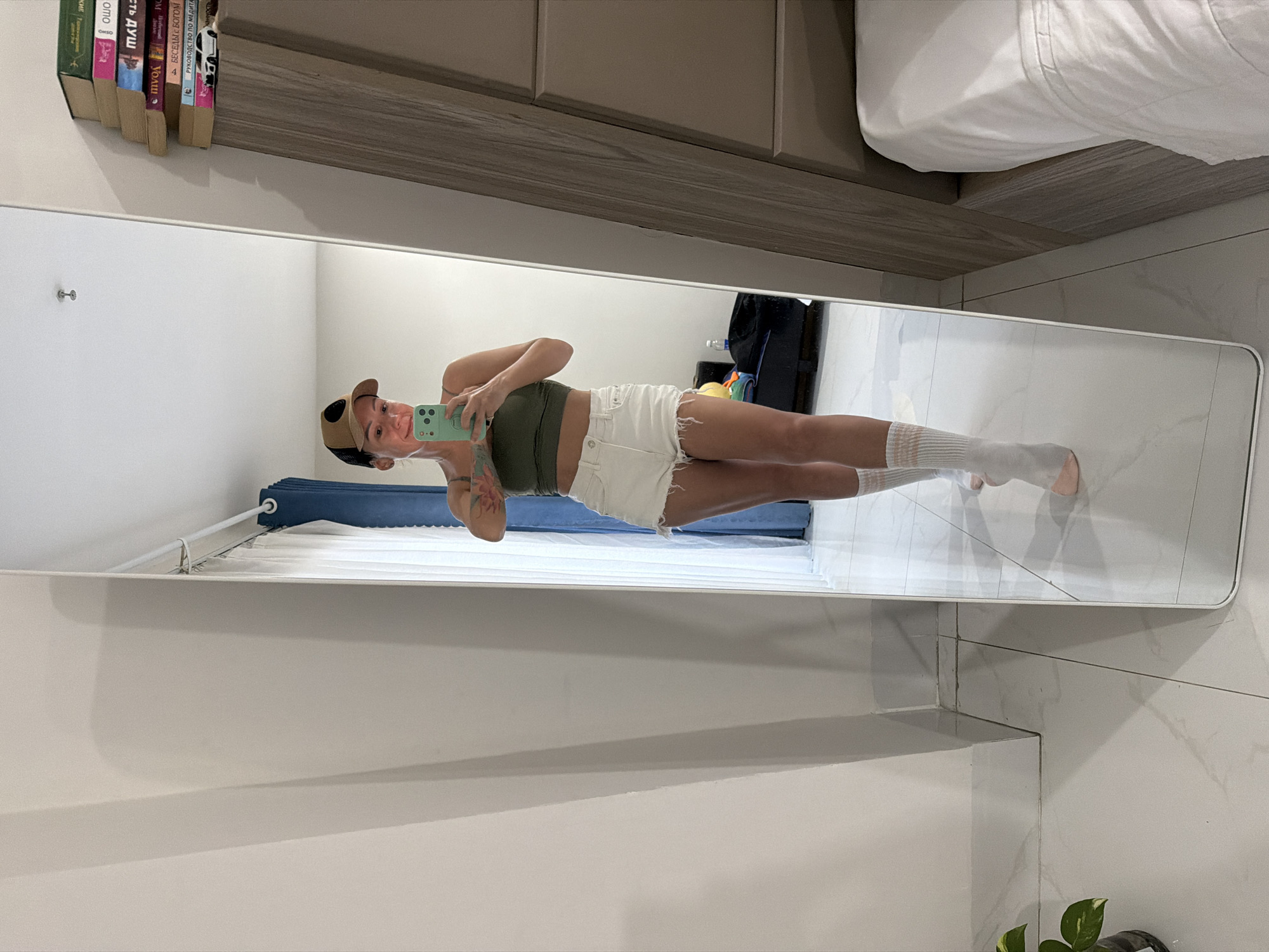 The image shows a full-length mirror reflecting a young woman standing in what appears to be a bedroom or living space. She is wearing a green crop top, white denim shorts, a beige baseball cap, and white knee-high socks. The woman is holding a mint green smartphone in her right hand, taking a selfie. She has tattoos visible on her arms.
The reflection reveals a bright, well-lit room with white tiled floors. In the background of the reflection, a bed with white linens is visible, and a wooden cabinet with books is mounted on the wall above. The cabinet holds several books with titles in Russian. A dark suitcase and a water bottle are also present in the reflection. The overall impression is casual and relaxed.
The image is taken indoors, and the lighting suggests it could be daytime. There are no explicit weather cues. The location context provided is Nha Trang, Vietnam, although there are no direct visual cues within the image to confirm this. The woman's expression is neutral and focused on taking the photograph.