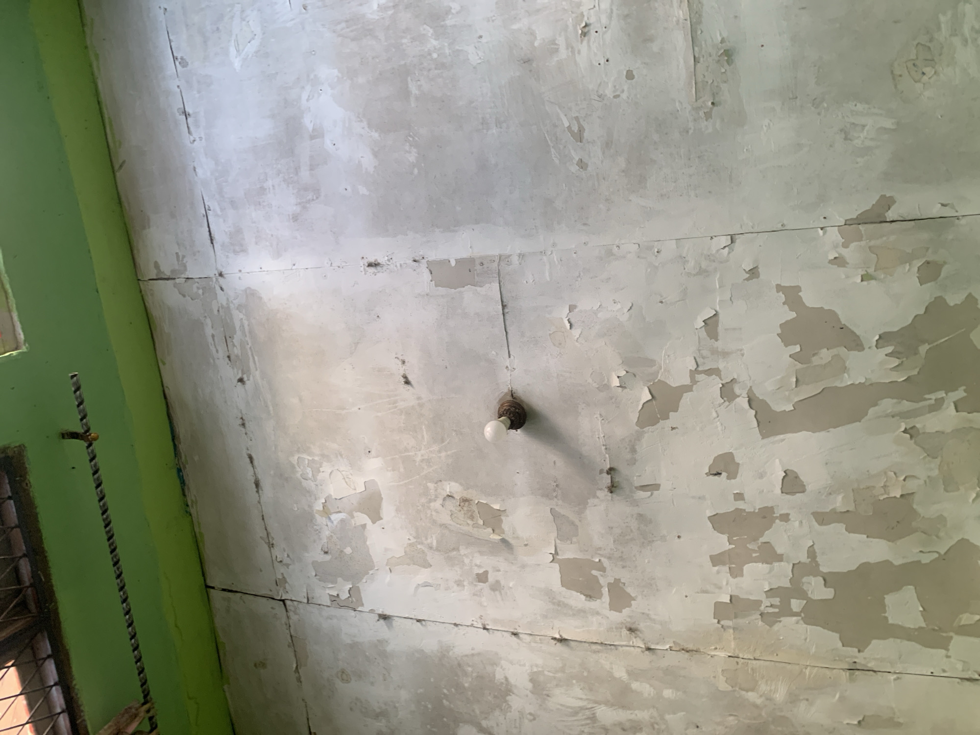 The image shows a cracked and peeling ceiling in what appears to be an interior space. A single light bulb is visible, mounted on a fixture attached to the ceiling. The walls to the left are painted bright green, and a metal grill is visible near the bottom left corner. The ceiling is composed of large panels, with visible seams and evidence of water damage or wear. The overall condition suggests neglect or a need for renovation. The image does not provide any specific information to indicate its location in Palembang, Indonesia.