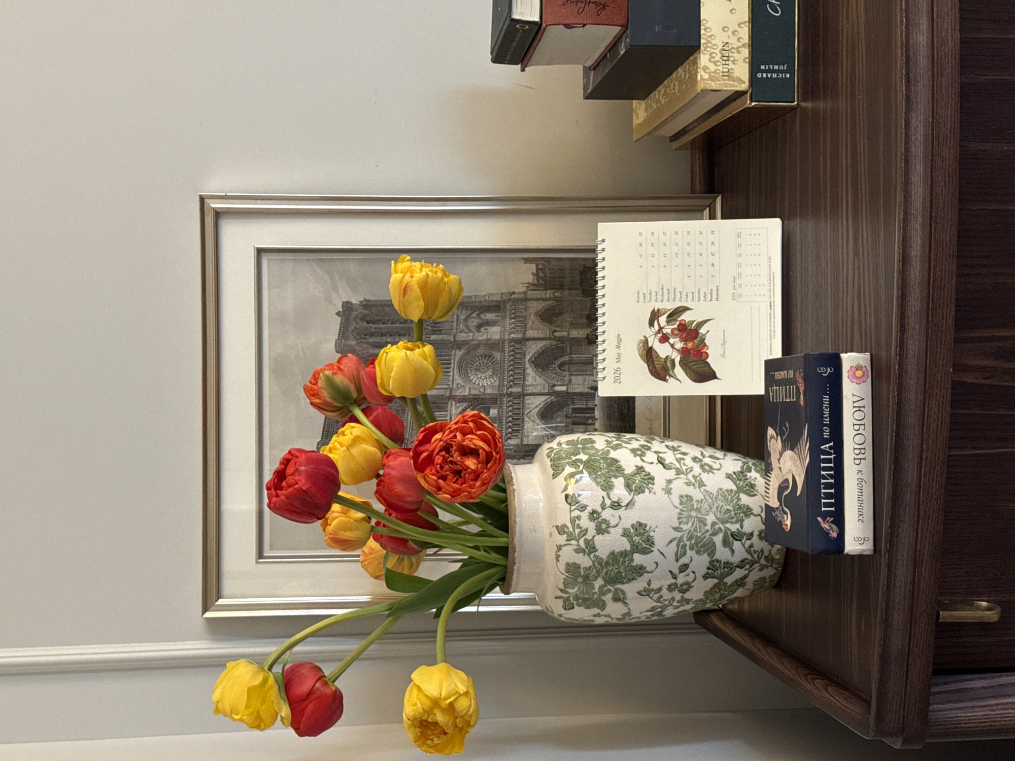 The scene is a still life arrangement. A bouquet of tulips in shades of red and yellow spills out of a white vase with a green floral pattern. Behind the vase is a framed black and white image, possibly of a church or cathedral. A calendar, showing "2026 May Maggio", rests partially on the frame and partially on a bookshelf. Two books are next to the vase on the bookshelf. The titles are partially visible and appear to be in Russian. The scene is set against a light grey wall. The location is a room in Moscow, Russia, inferred from the Russian titles of the books.