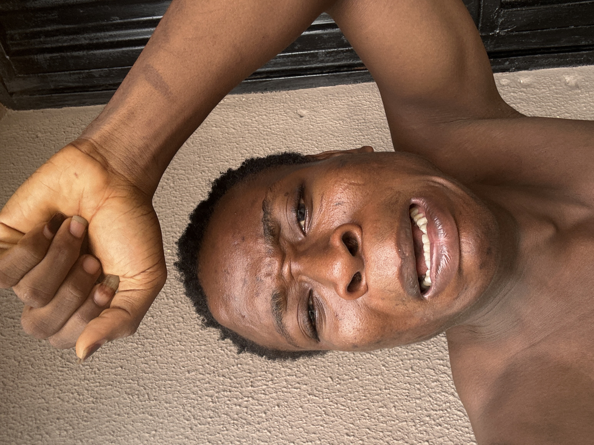 A close-up, low-angle shot shows a dark-skinned person lying on a beige carpet, with a black window frame or similar structure visible in the upper left corner. The person's face is centered in the frame, displaying a contorted expression with eyes squinted and lips parted to reveal teeth. One arm is raised, with the hand positioned near the side of the face. The overall impression is one of discomfort or duress. The location is unknown.