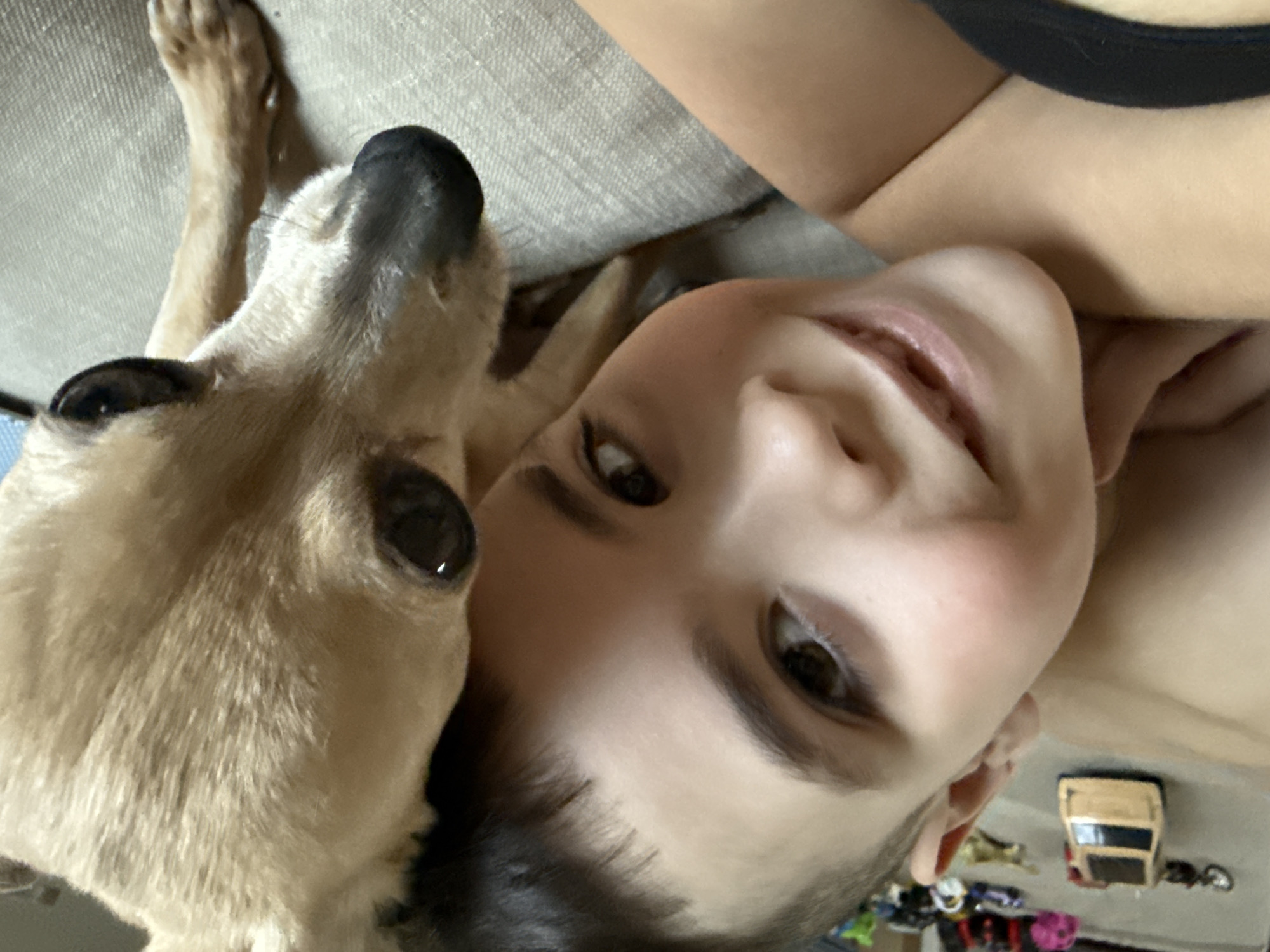 An indoor, close-up photograph taken in Chaikovsky, Russia, depicts a young child and a small dog. The child, with fair skin, short dark hair, and dark eyes, is positioned on the left, smiling directly at the camera. A black garment strap is visible near the child's lower left. To the child's right, a tan-furred dog, likely a Chihuahua, with dark eyes and a black nose, is partially visible, positioned close to the child. Both subjects are situated on a light-colored, textured fabric surface, possibly a couch or bed. In the blurred background, several small toys are present, including a prominent tan toy car.
