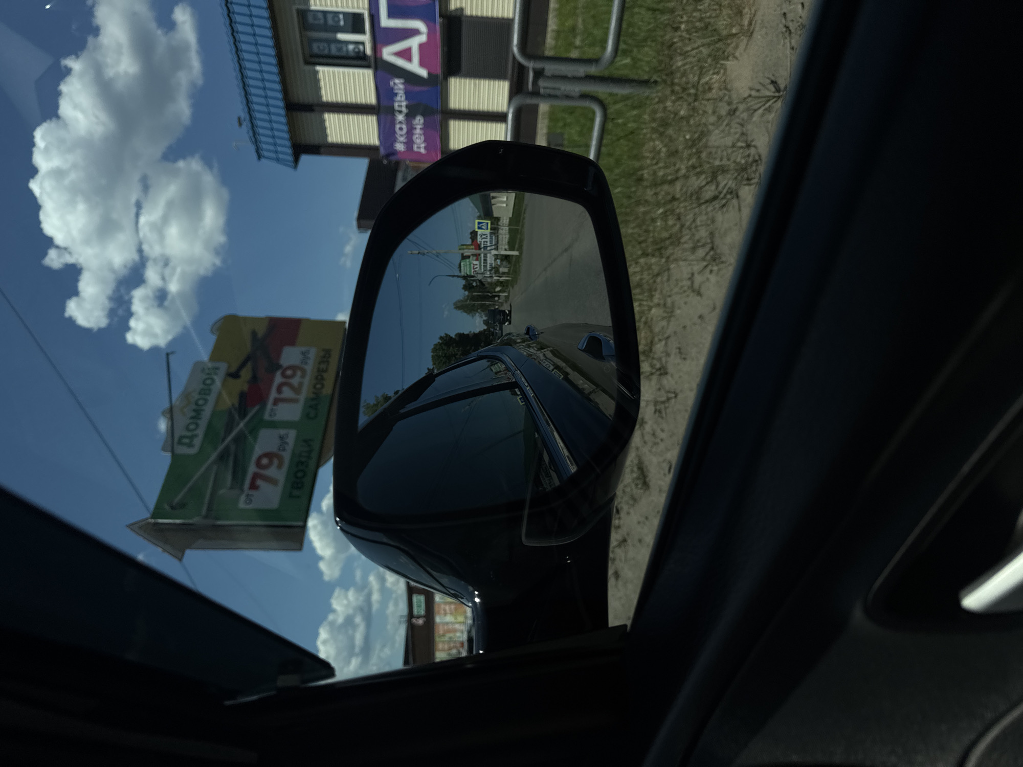 The image shows a view from inside a car looking out the passenger window and into the side mirror. The car appears to be on a road in Chaikovsky, Russia, as indicated by the sign in the distance.
In the foreground, the dark interior of the car is visible, including the door panel and a portion of the window frame. The side mirror, reflecting the road and the sky, is a prominent feature.
Outside the car, a street scene unfolds. A large green and yellow billboard with Russian text and prices is visible on the left side of the road. The text on the billboard appears to advertise building materials, with prices like "79 rub." and "129 rub." and words like "гвозди" (nails) and "саморезы" (screws).
Further down the road, a two-story building with a blue roof and a facade of light-colored horizontal siding is present. A banner with "#каждый" (every) and the letters "АГ" is displayed on this building. Other buildings and signs are visible in the background, including a sign that reads "ЗАПЧАСТИ" (spare parts) and a green triangular sign with an 'A' inside, possibly a road sign.
The sky is a bright blue with scattered white clouds. The ground alongside the road appears to be covered with dry grass and sand. A metal railing is visible in the middle ground. The overall scene suggests a sunny day in a semi-urban or suburban area.