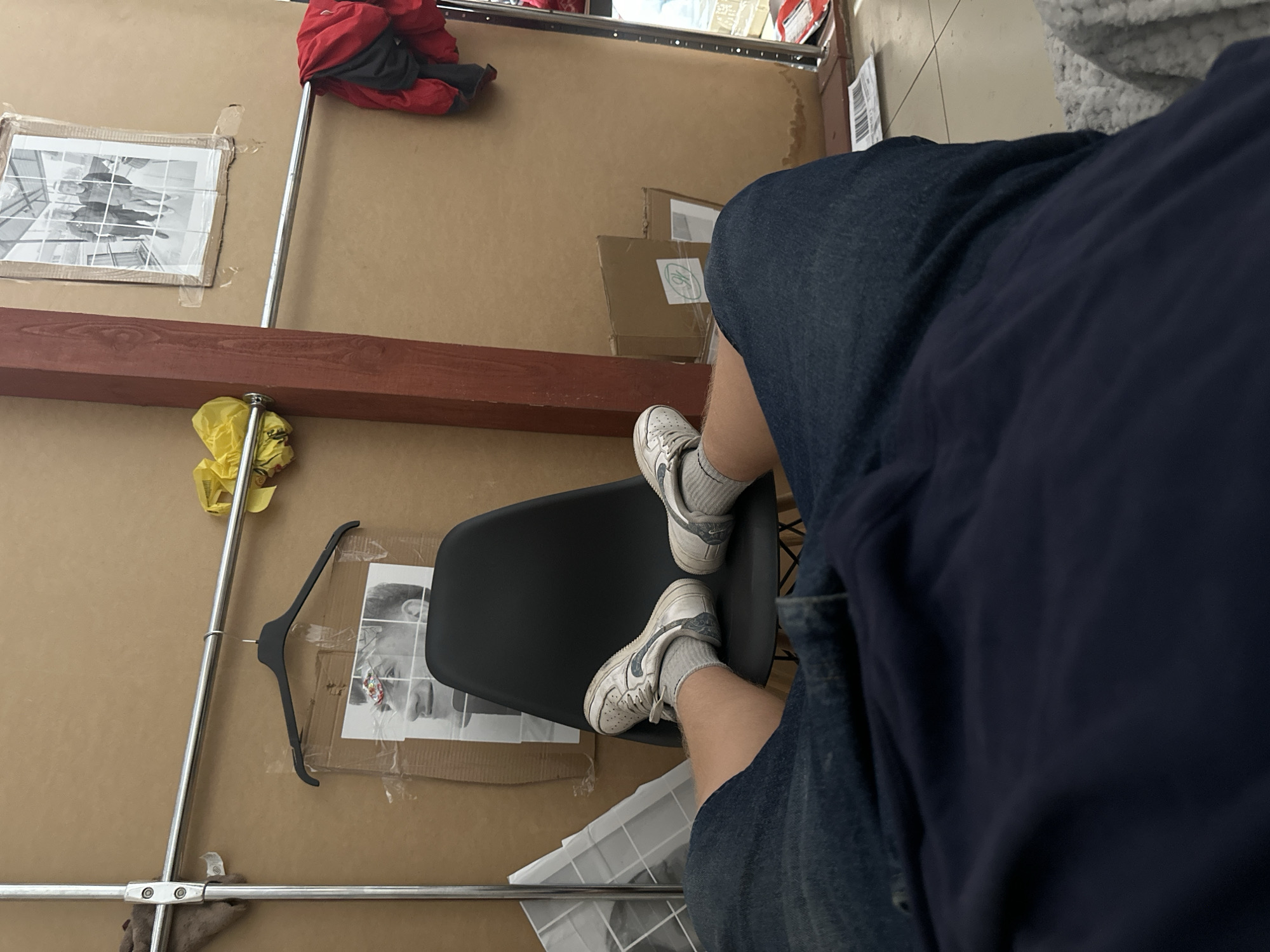 The image shows a person's legs and feet resting on a black chair. The person is wearing blue denim shorts and white sneakers with grey socks. The background consists of a wall with some framed pictures and a coat rack with clothes hanging on it. There are also some boxes and a yellow bag visible. The scene appears to be indoors, possibly a room or a storage area. The location in Khmilnyk, Ukraine is not discernible from the image itself.