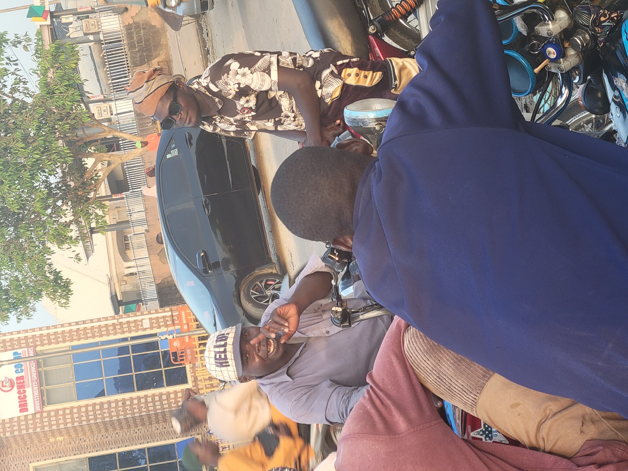 The image shows a group of men interacting on a street in Madalla, Nigeria. A man wearing a floral shirt, traditional cap, and sunglasses stands next to a black car and appears to be talking to others. Two men are sitting on a motorcycle, one wearing a "HELLO" cap and gesturing with his hand. A third man in a blue jacket is partially visible, with the focus on the back of his head and shoulder. In the background is a building with a sign that reads "DRICCHER CO OP". The street scene is captured in daylight, suggesting it is a sunny day.