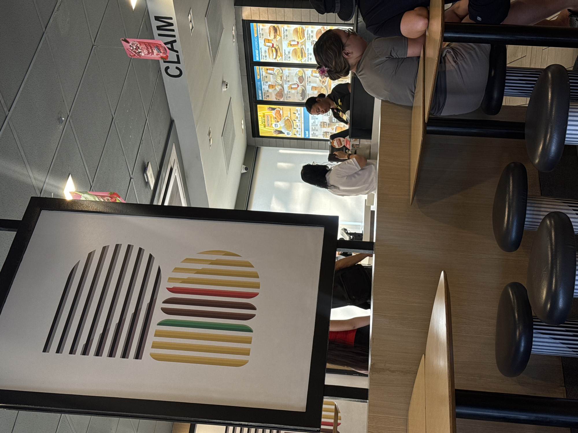 The image captures the interior of a fast-food restaurant, likely a McDonald's, in Davao City, Philippines. The perspective is from a low angle, looking towards a service counter and seating area.
In the foreground and mid-ground, a long, light-colored wooden counter extends horizontally, bordered by black-framed artwork depicting abstract, striped burger elements in various colors (brown, red, green, yellow, white, black). Along this counter are several black, oval-shaped padded stools, some occupied.
To the right, a person with dark hair tied back with a pink flower clip, wearing a grey shirt, is seated at the counter, facing away from the camera. Next to them, parts of another person in dark clothing are visible.
At the service counter further back, a staff member in a dark uniform with yellow accents is attending to a customer. This customer, with dark hair and wearing a white shirt, appears to be ordering or waiting for food. Above the service counter, large digital menu boards display various food items, including "BREAKFAST SANDWICHES," "BREAKFAST PLATTERS," "BURGER & RICE MEALS," and "BEEF," with prices in Philippine Pesos (e.g., "P129," "P184").
Overhead, a "CLAIM" sign in black lettering on a white background hangs from the ceiling. A pink promotional banner, possibly for a dessert item like McFlurry, also hangs nearby. The ceiling features grey acoustic tiles with embedded lighting fixtures. The overall ambiance suggests a bright, modern fast-food setting during daytime.