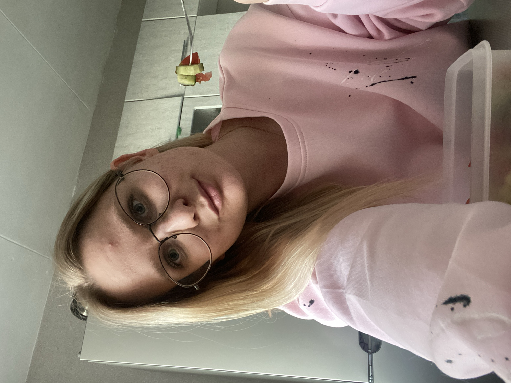 A fair-skinned woman with blonde hair and round eyeglasses is the primary subject of the image. She is wearing a pink sweatshirt with what appears to be paint splatters on the front. She is holding a clear plastic container in her right hand. A silver fork with food speared on it is suspended in the upper left of the image. The background is a neutral-colored tiled wall and a metallic surface, likely a kitchen or bathroom.