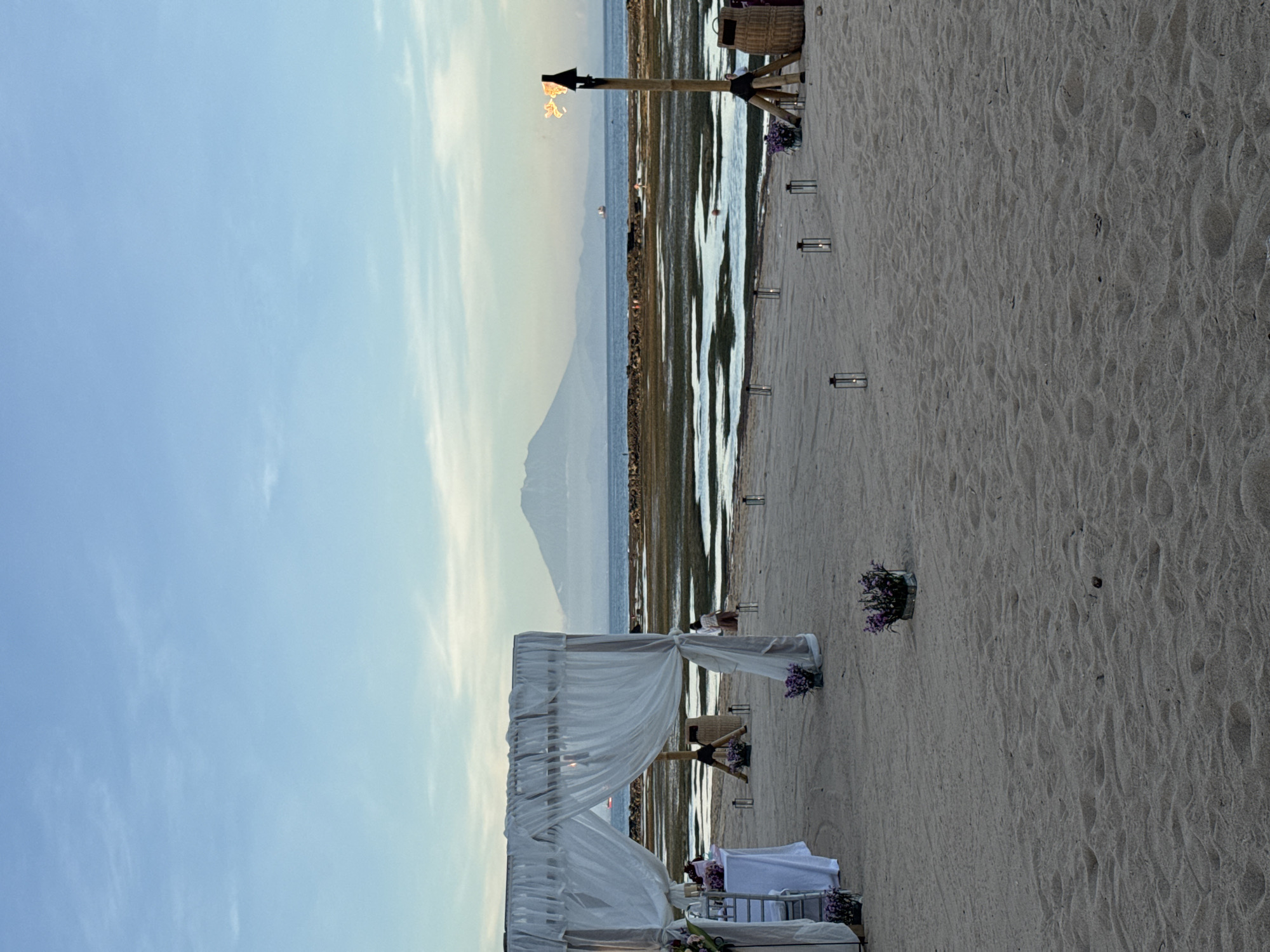 The image depicts a beach setting, presumably for a wedding, as indicated by the white-draped wedding altar positioned in the foreground. The scene is set at the water's edge, with shallow water revealing the seabed. A mountain is visible in the background, partially obscured by the sky. Several small lanterns are spaced along the beach, and a torch burns on a wooden stand. No people are clearly visible in the image. The location is the city of Unknown, Unknown.