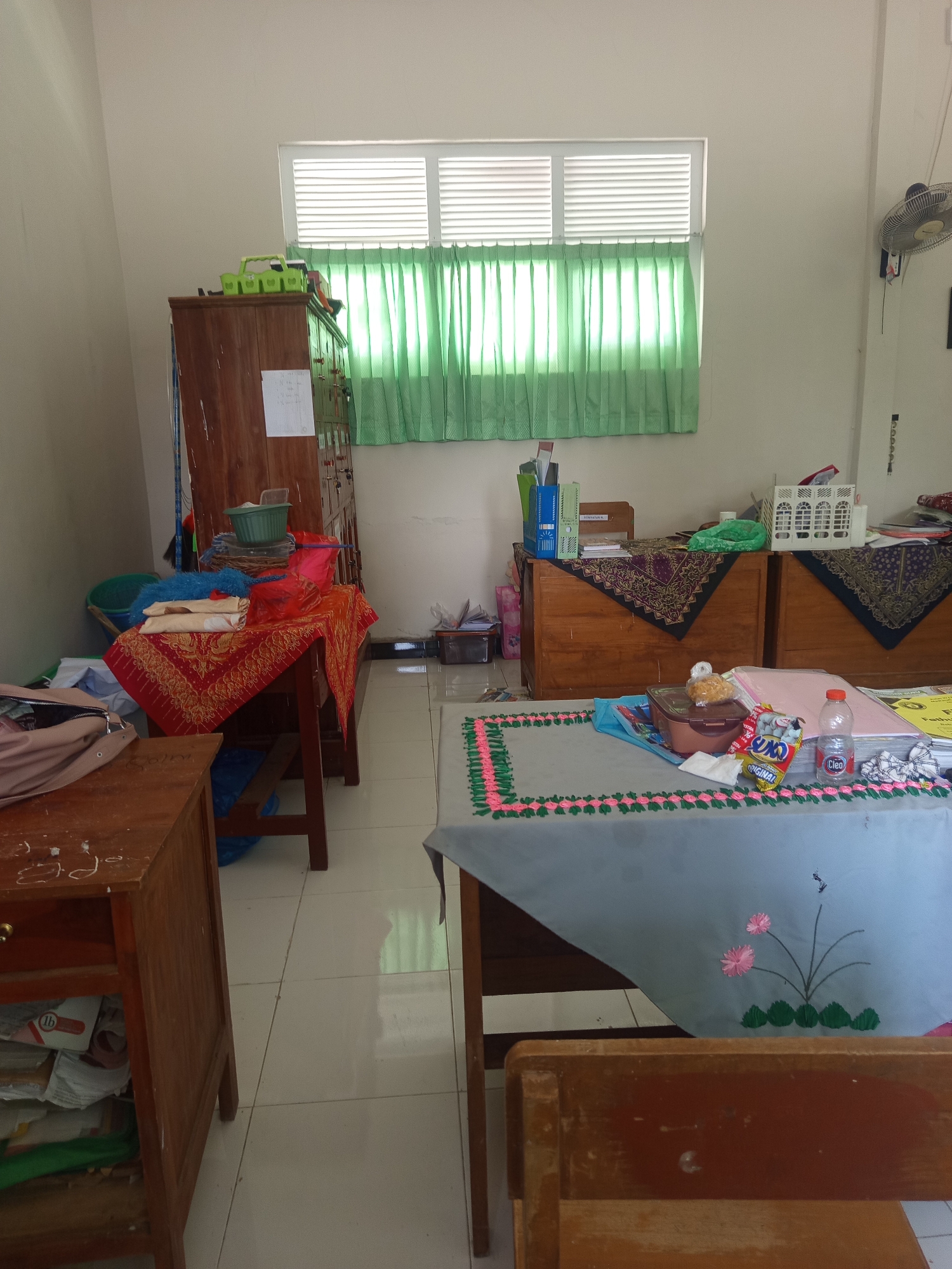 The image depicts a classroom or office space in Lumutan, Indonesia. The room is filled with various items, suggesting a functional but somewhat cluttered environment. In the foreground, a wooden desk with drawers is visible on the left, containing stacks of paper and some plastic bags. To the right, another wooden desk is partially visible, occupied by a wooden chair.
A prominent feature of the room is a large wooden cabinet to the left of center, displaying several items on its top, including a green plastic container and some stacked objects. In front of the cabinet, a smaller wooden table is covered with a vibrant red and gold patterned cloth, on which more items are arranged, such as folded fabrics, a blue feather duster, and a green plastic basket.
The background reveals a window covered with light green curtains that let in diffused sunlight. To the right of the window, a wall-mounted fan is visible. Further to the right, two large wooden cabinets or chests of drawers are placed side-by-side. These are draped with patterned cloths, and on top, various items including binders, boxes, and a white plastic crate are present.
In the middle ground, a long table is covered with a light gray cloth that has a decorative border and embroidered flowers. On this table, there are food items in packaging, a bottle of "Cleo" water, and other miscellaneous objects. The floor is tiled with light-colored square tiles. The overall impression is of a lived-in space, possibly a teacher's office or a classroom storage area, with evidence of daily use and personal belongings.