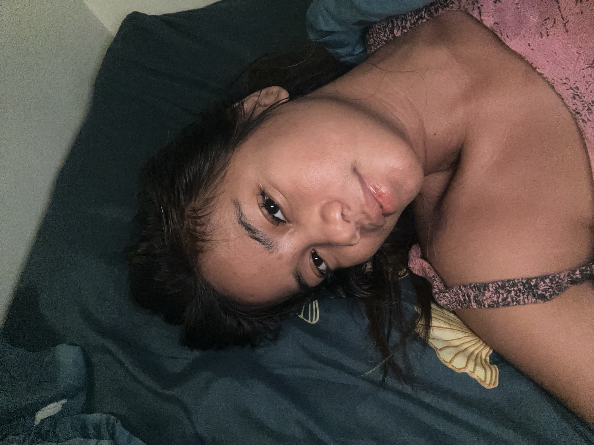 An overhead, slightly angled shot captures a young person lying on a dark teal bedspread. The person is on their right side, facing the camera, with their head tilted. They have dark hair that falls across their shoulder and face. Their skin is a warm tone, and they are wearing a pink top with a patterned trim. The background consists of the bedspread and a hint of a light-colored wall on the left. The overall lighting suggests an indoor setting, possibly a bedroom. The image does not contain any specific indicators of the city of Sukawati, Indonesia.