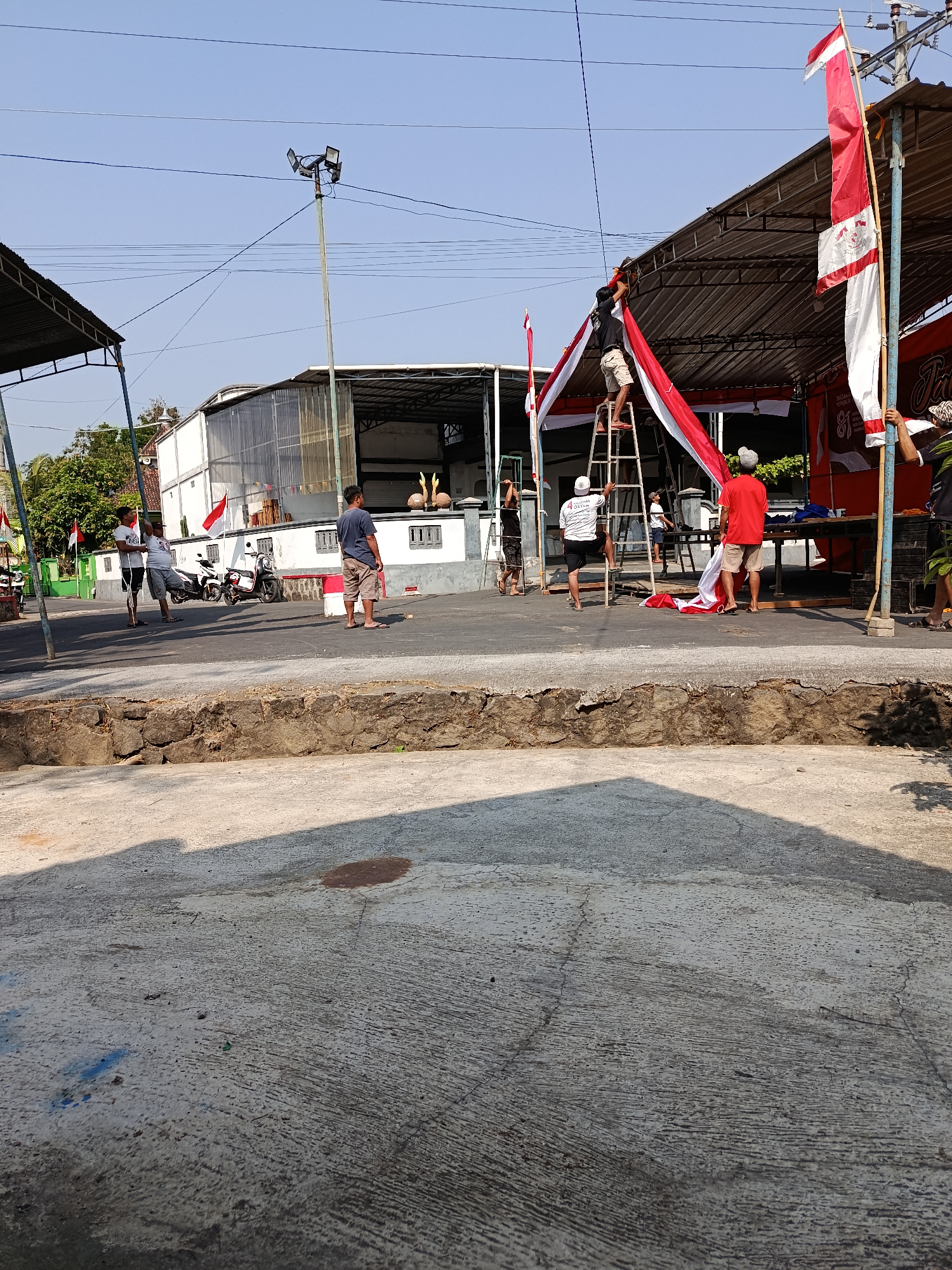 In Jurug, Indonesia, a group of individuals are actively preparing an outdoor event space, possibly for a celebration. The scene is dominated by the installation of red and white banners, reflecting the Indonesian national colors. Several people are engaged in this task, with some on ladders securing the fabric to a temporary canopy structure. One man in a red shirt is holding a length of banner, while another is on a ladder, attaching it. A third person, wearing a white shirt with the number "4" on it, is also on a ladder, assisting with the decorations.
The environment suggests a sunny day with a clear blue sky. In the background, there are several buildings, including one with a corrugated metal roof and another that appears to be a more modern structure with glass panels. Motorcycles are parked nearby, indicating the presence of transportation. A tall pole with multiple lights is also visible, likely for evening illumination. The foreground shows a concrete surface, with a raised stone wall and some shadowing from the overhead structures and the sun. The overall impression is one of community effort and preparation for an upcoming festive occasion.
