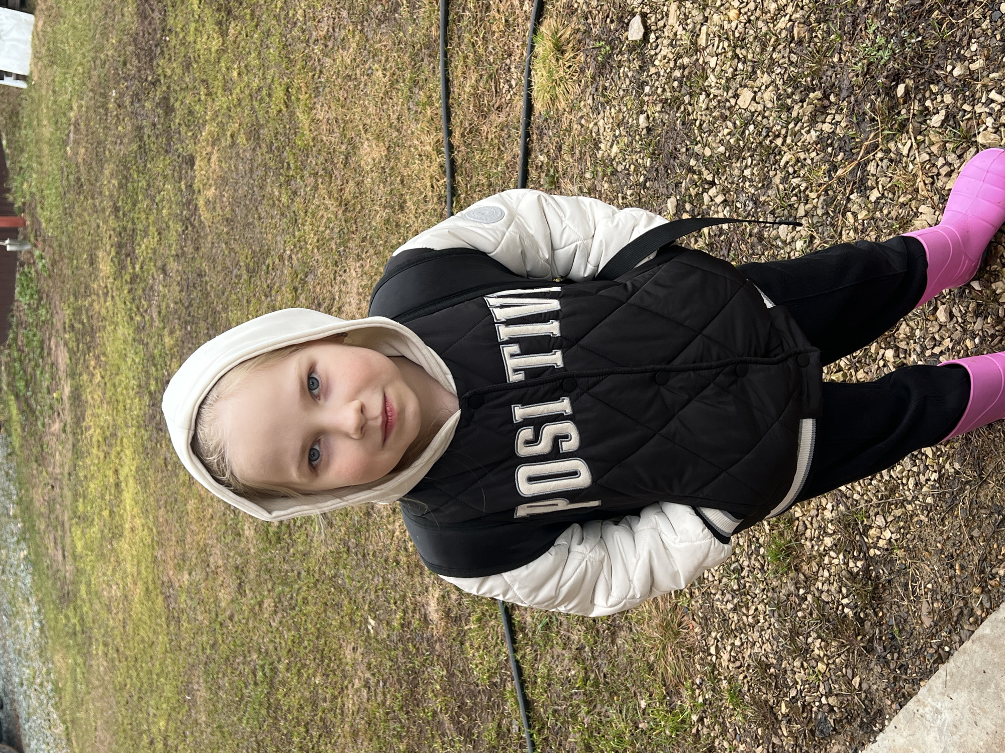 A young child with fair skin, blue eyes, and blonde hair stands outdoors. The child wears a white hooded jacket with a black quilted vest over it, the vest featuring the word "POSITIVE" in white block letters. The child also wears black pants and bright pink rubber boots. The ground consists of grass and gravel. The location is Bol'shakino, Russia.