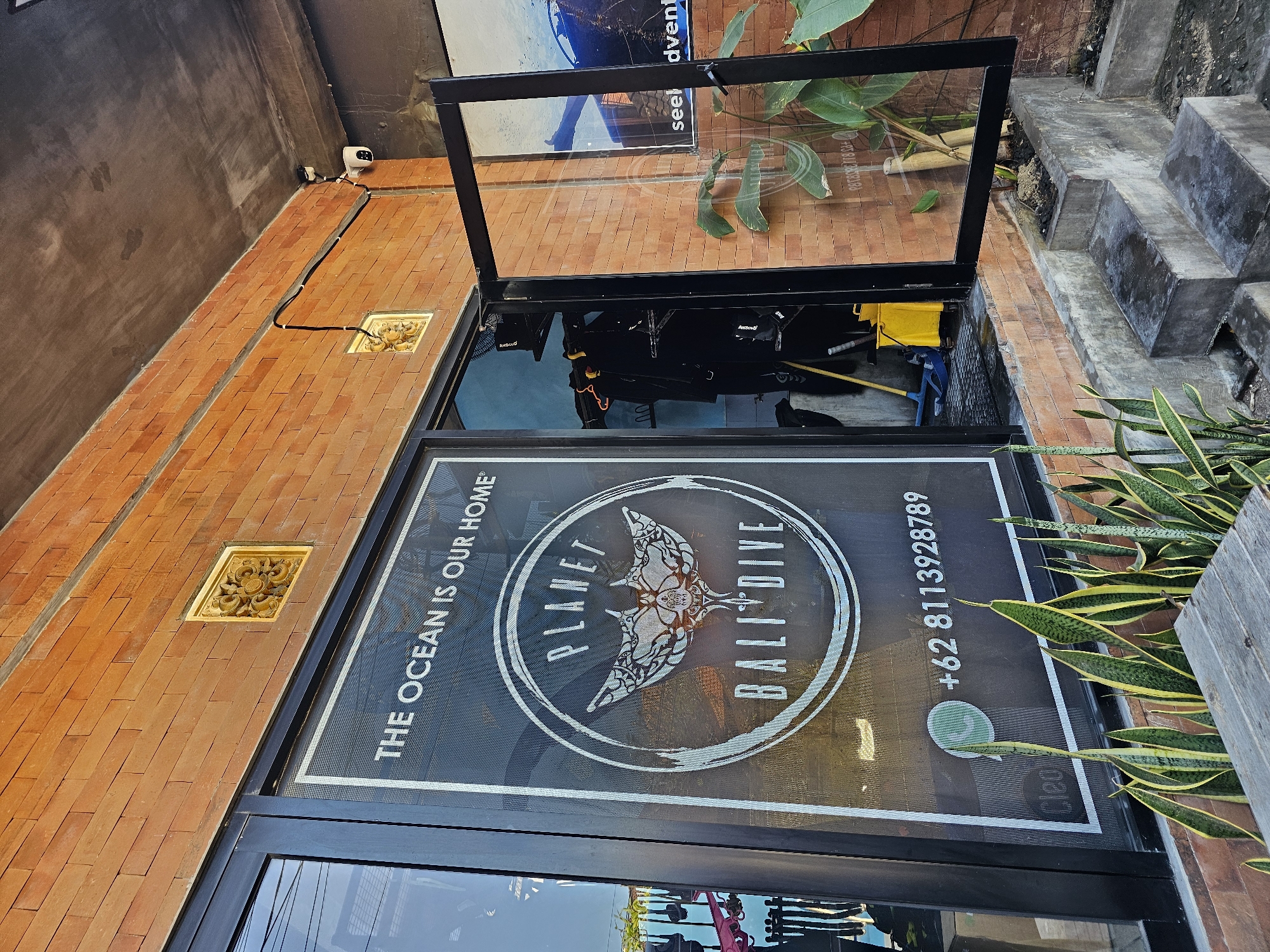 This is a medium-shot, eye-level, exterior, daytime, photograph of the entrance to "Planet Bali Dive" located in Denpasar, Indonesia. The building facade is constructed of warm-toned brick. A large glass storefront window dominates the left side of the frame, featuring a white graphic of a manta ray with the text "PLANET BALI DIVE" and "THE OCEAN IS OUR HOME". Below the graphic, a white phone number, "+62 8113928789", is displayed. To the right of the window, a glass door is ajar, revealing diving equipment, including wetsuits and a fan, inside. The exterior also features a security camera mounted on the wall, decorative golden wall ornaments, and a planter with snake plants. Concrete steps lead up to the entrance. The overall scene depicts a dive shop entrance in a tropical environment.