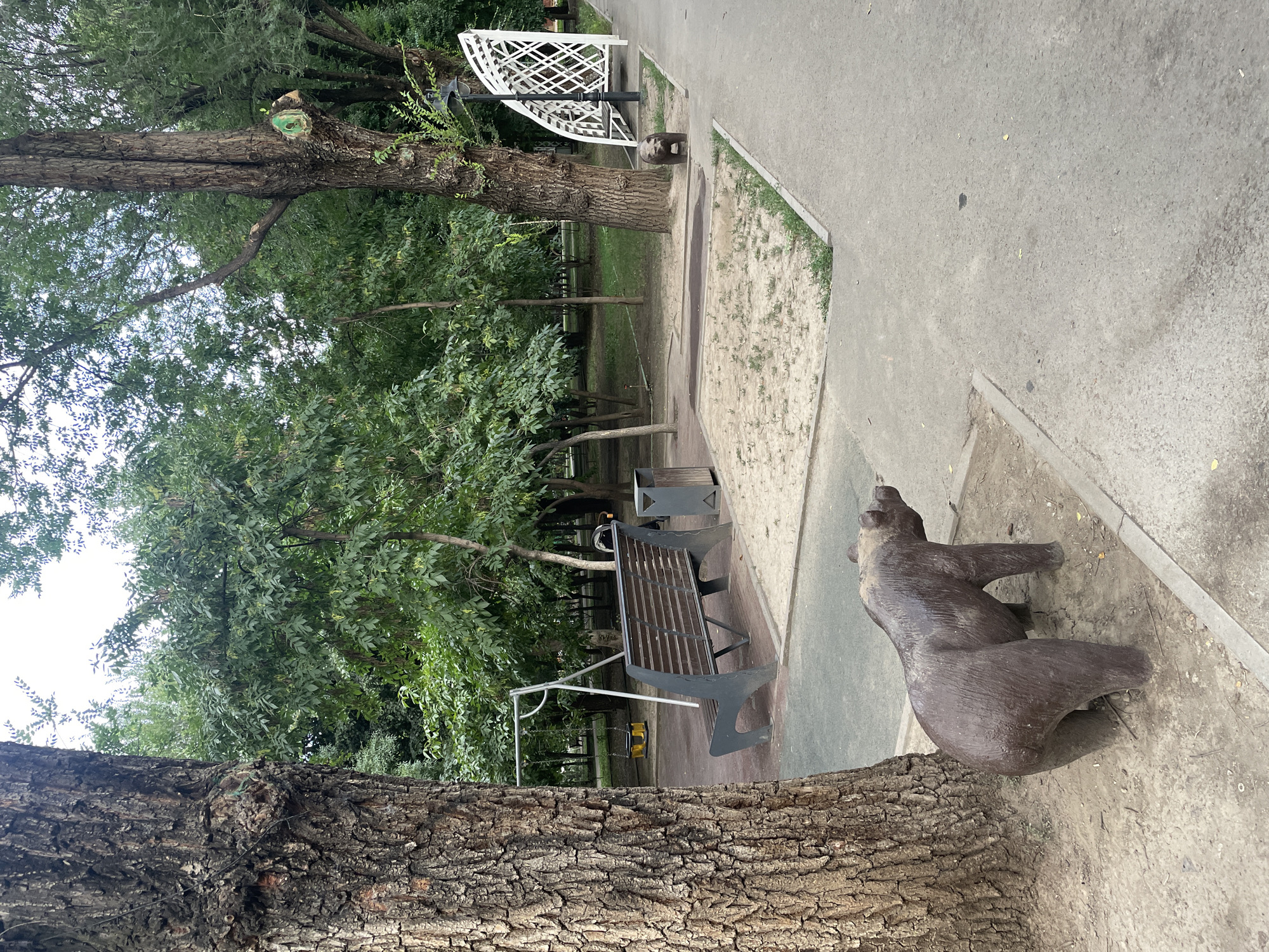 The image depicts a park scene in Almaty, Kazakhstan, featuring a concrete path winding through a grassy area with numerous trees. In the foreground, a large tree trunk is prominent on the left. A brown bear sculpture stands near the base of another tree on the right side of the path. Further into the park, a metal swing set with a yellow seat is visible, along with a wooden bench and a dark grey trash receptacle. A white decorative arch structure stands behind the bench and trash can. The ground in the park consists of sand and dirt, with some patches of grass. The overall atmosphere is that of a public park during the daytime.