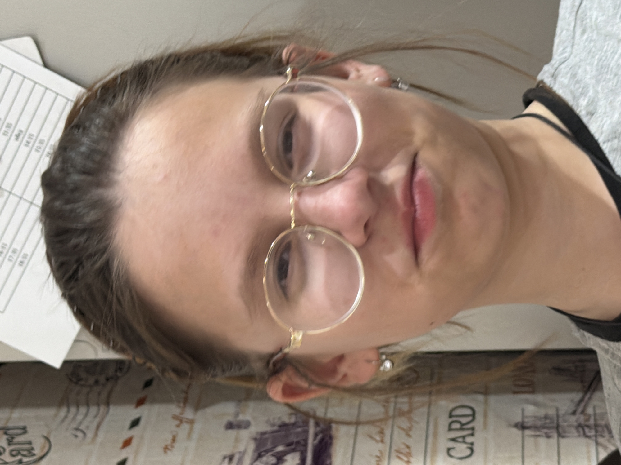A close-up, tilted shot of a woman wearing round, gold-rimmed glasses. She has light skin and brown hair. She is wearing a grey t-shirt with a black tank top underneath. To the left, there is a piece of paper with times written on it and a postcard-themed surface visible. The image does not provide information about the location in the city of Chaikovsky, Russia.