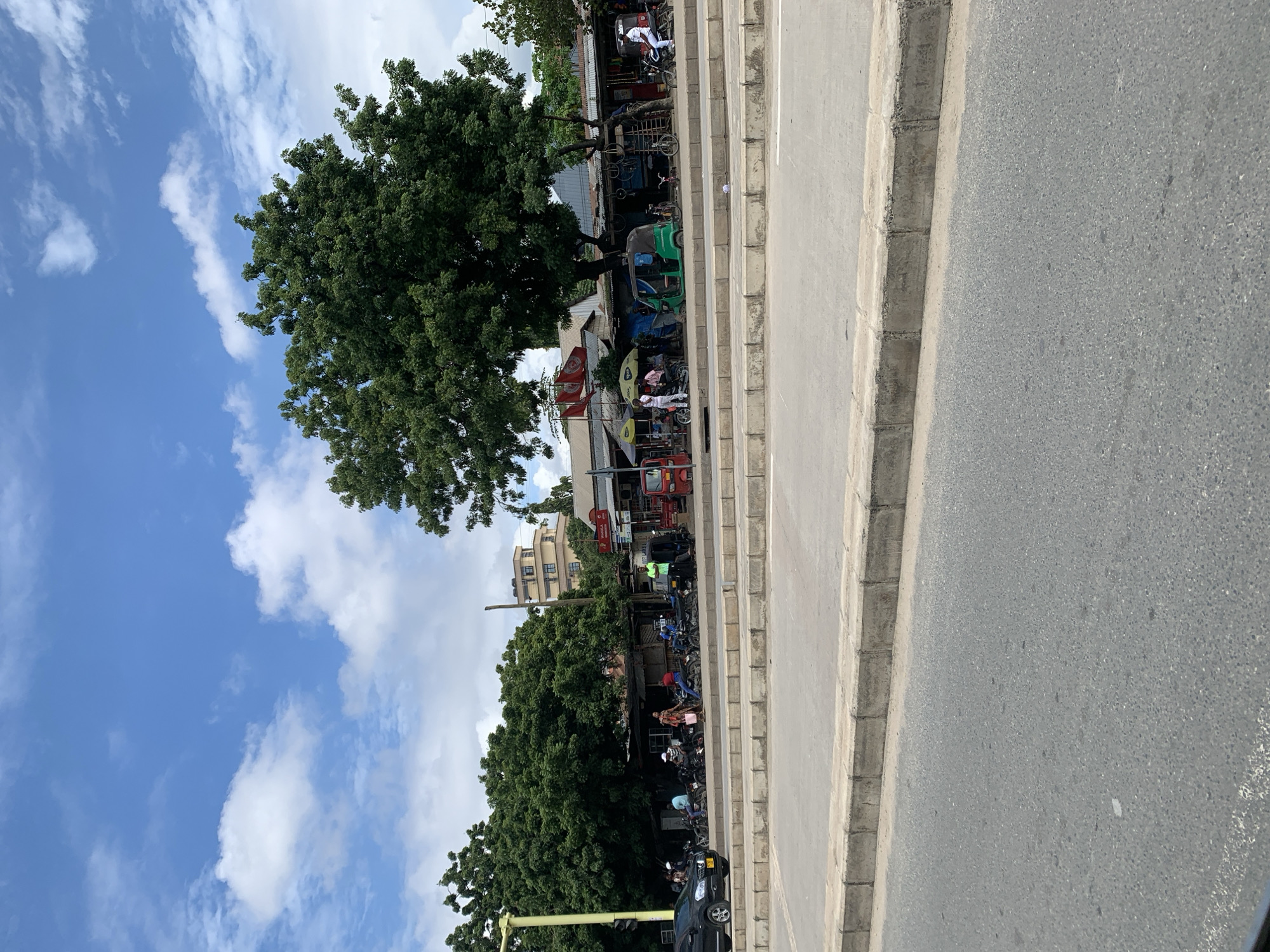 The image depicts a street scene in Dar es-Salaam, Tanzania. The foreground consists of a paved road and a concrete curb. Along the side of the road, there are shops and businesses shaded by trees. There are motorcycles and auto rickshaws parked alongside the shops. People can be seen standing near the shops. In the background, a building is visible. The sky is blue with scattered clouds.