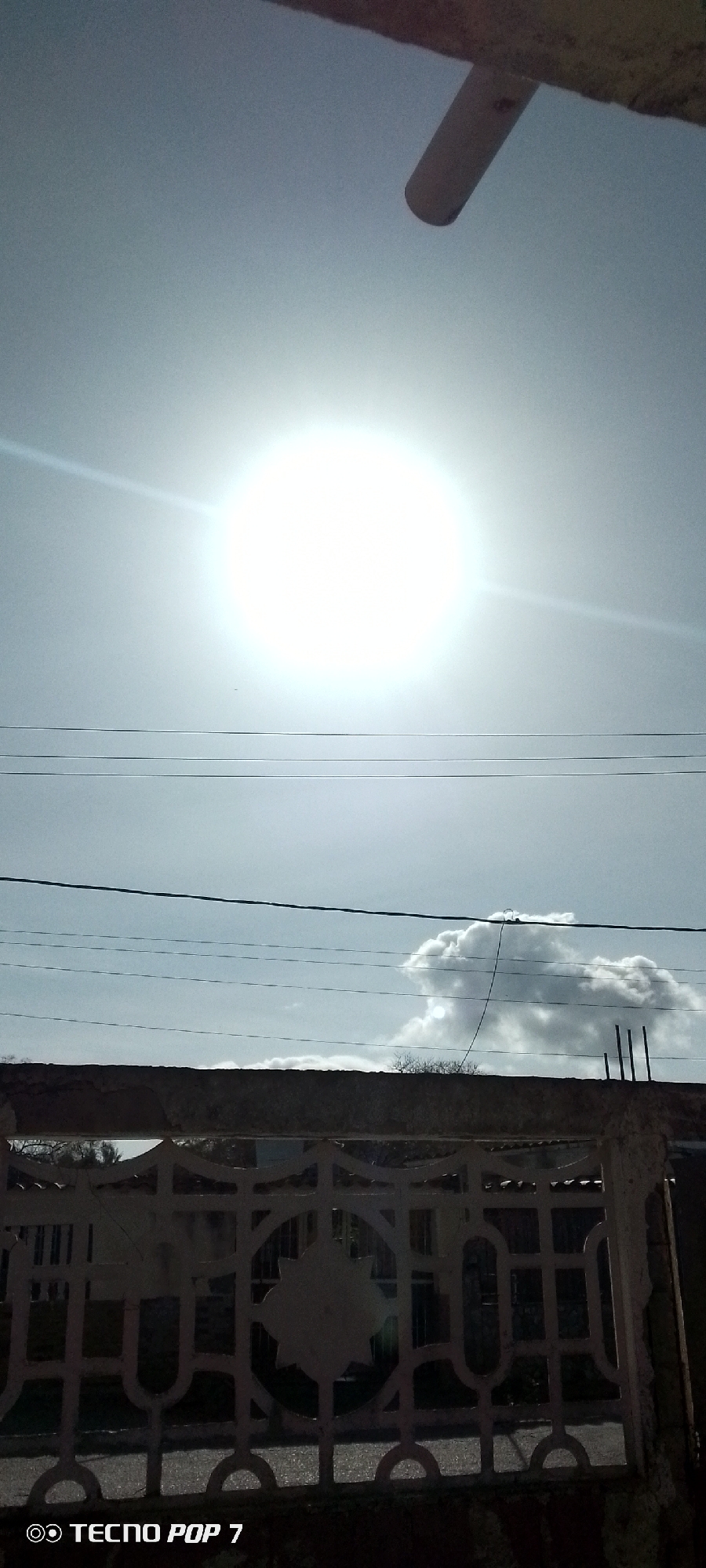 The image shows a bright, sunlit sky in Altagracia, Venezuela. The sun is a large, bright orb in the upper half of the frame. Below the sun, there are a few power lines stretching across the view. Further down, a thick cloud formation is visible. In the foreground, a decorative fence partially obscures the view of a building behind it. A portion of what appears to be a ceiling fan or light fixture is visible at the top of the image. "TECNO POP 7" is printed at the bottom left corner.