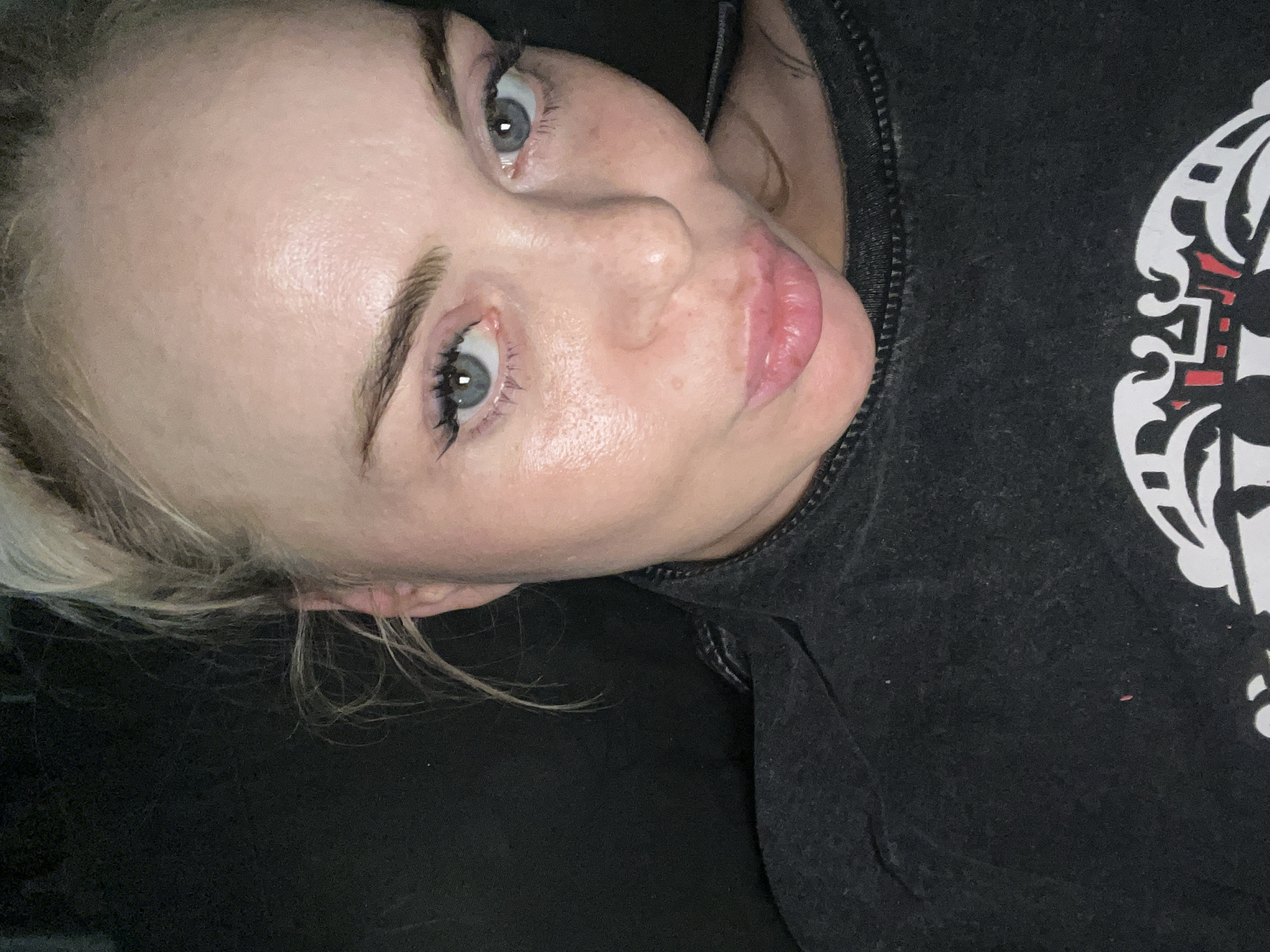 A close-up, high-angle selfie shows a blonde woman with blue eyes and pink lips, wearing a dark t-shirt, against a dark background. The woman's hair is pulled up in a messy bun, with strands of hair falling around her face. Her face is turned slightly to the left, and she is looking at the camera with a neutral expression. The lighting is dim, with a strong light source coming from the front, creating highlights on her skin. There appears to be some smudging on the t-shirt. The background is a dark, out-of-focus area.