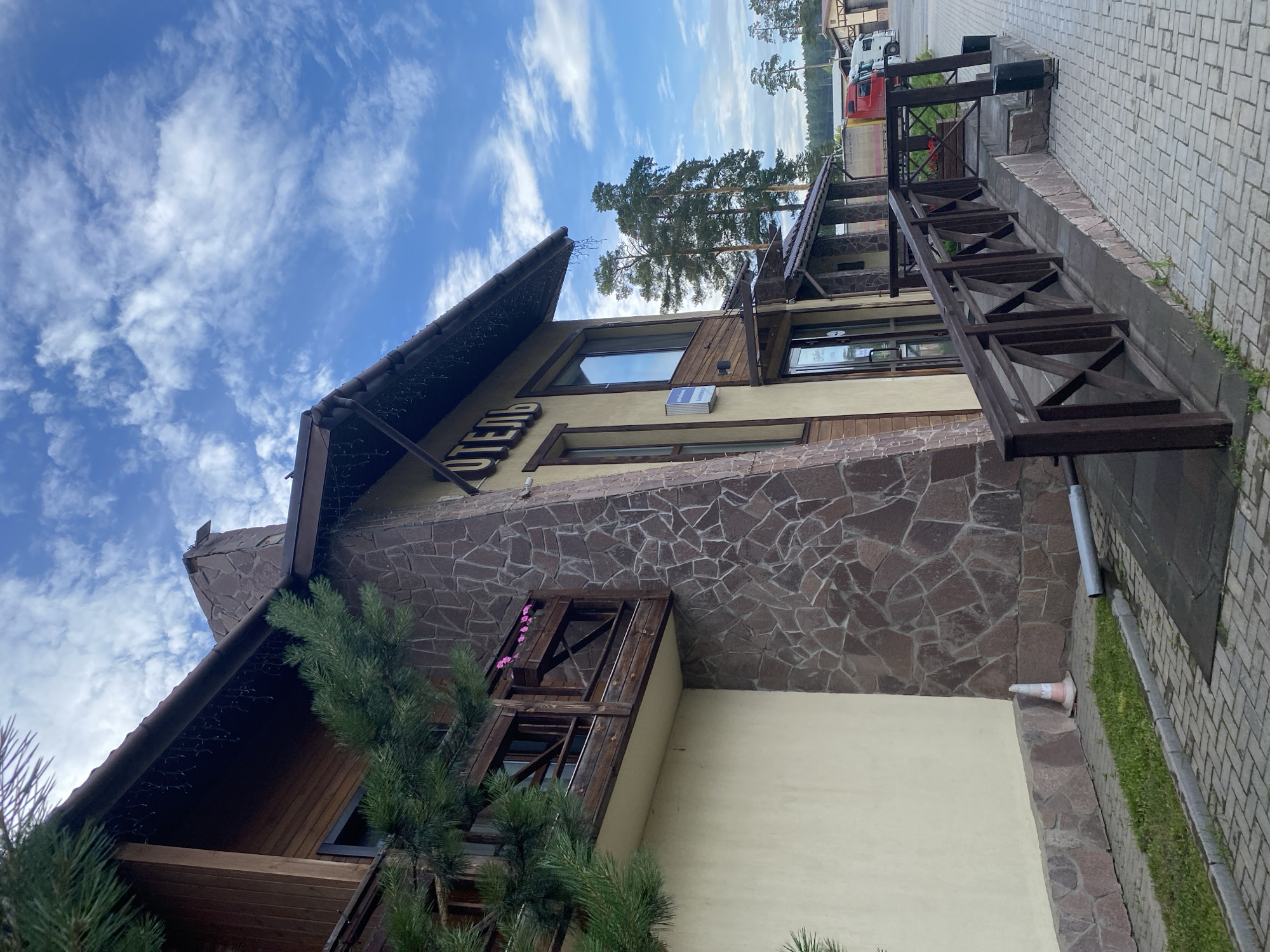 This media file captures a low-angle view of a hotel building with a stone and wood facade under a bright, partly cloudy sky. The building has a steeply pitched roof adorned with decorative lights, and a wooden porch or balcony area. A dark wooden railing with a geometric pattern runs along an elevated walkway.
The building has large windows, some with wooden frames. A sign with Cyrillic text, "ОТЕЛЬ" (Hotel), is visible on the upper section of the building. Pink flowers are placed in a wooden planter box above one of the windows. To the right of the building, a paved walkway with a textured brick pattern leads away, bordered by a strip of grass. A traffic cone is visible near the base of the building.
In the background, tall pine trees are silhouetted against the sky. A red and white vehicle, possibly a truck or van, is parked further down the paved area, suggesting it's a road leading to or from the hotel. The presence of the vehicle and the clear sky with scattered clouds indicates it is likely daytime. The overall scene conveys a sense of a welcoming, possibly rustic, lodging establishment in a natural, wooded setting, likely Votkinsk, Russia.