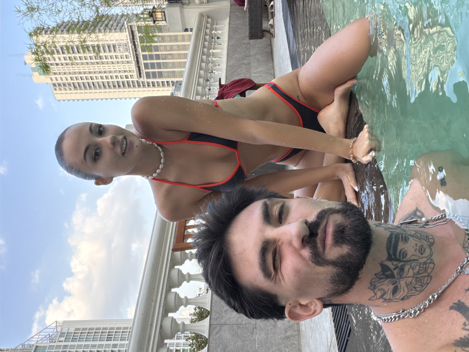 A daytime image depicts two individuals in a rooftop pool setting, located in Bangkok, Thailand, identifiable by the prominent "SIAM" signage and partially visible "HOTEL" text on a distant high-rise building. In the foreground, a male subject is partially submerged in light blue-green water, facing the camera with his left eye closed in a wink. He possesses dark hair, a beard, and a mustache. Multiple silver-colored chain necklaces are worn around his neck, displaying prominent tattoos depicting stylized faces. To his right, a female subject is positioned kneeling on a wet surface at the pool's edge, with her lower legs submerged in the water. She has dark hair pulled back, and water droplets are visible on her skin. She wears a dark-colored bikini with red trim, a white cowrie shell necklace, and a light-colored beaded bracelet on her left wrist, looking towards the camera with a slight smile. The background features a white classical-style balustrade, a wooden chair, and several tall urban buildings under a partially cloudy blue sky. Green foliage is visible on the far right, and a dark-colored lounge chair with a red towel is present further back.