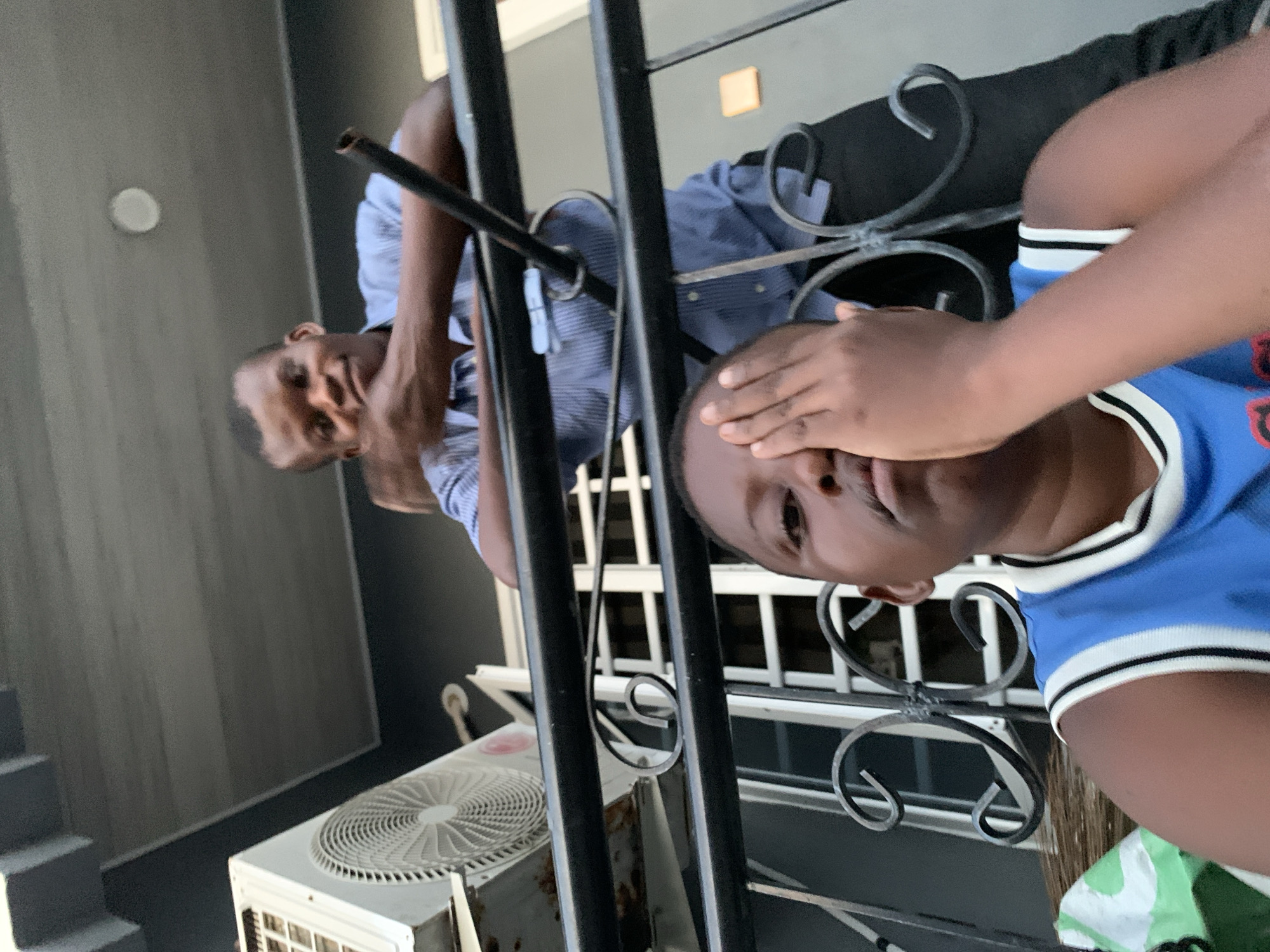 The image, taken in Ibasa, Nigeria, features two young males in an outdoor or semi-outdoor setting, separated by decorative black metal railings. In the foreground, a male wearing a blue and white striped short-sleeved top has his right hand covering the upper part of his face, looking towards the camera. Behind the railings, another male, dressed in a light blue striped collared shirt and black trousers, is partially visible, looking directly at the camera with a neutral to slightly smiling expression.
In the background, to the left, grey steps ascend alongside a grey wall. An outdoor air conditioning unit with a circular fan vent is situated on the ground in the lower-left corner. To the right, a darker wall section features a small, light-colored square object, possibly a light fixture. The scene is brightly lit, indicating daytime, possibly with soft or indirect sunlight. The overall atmosphere appears casual. No visible text is present in the image.