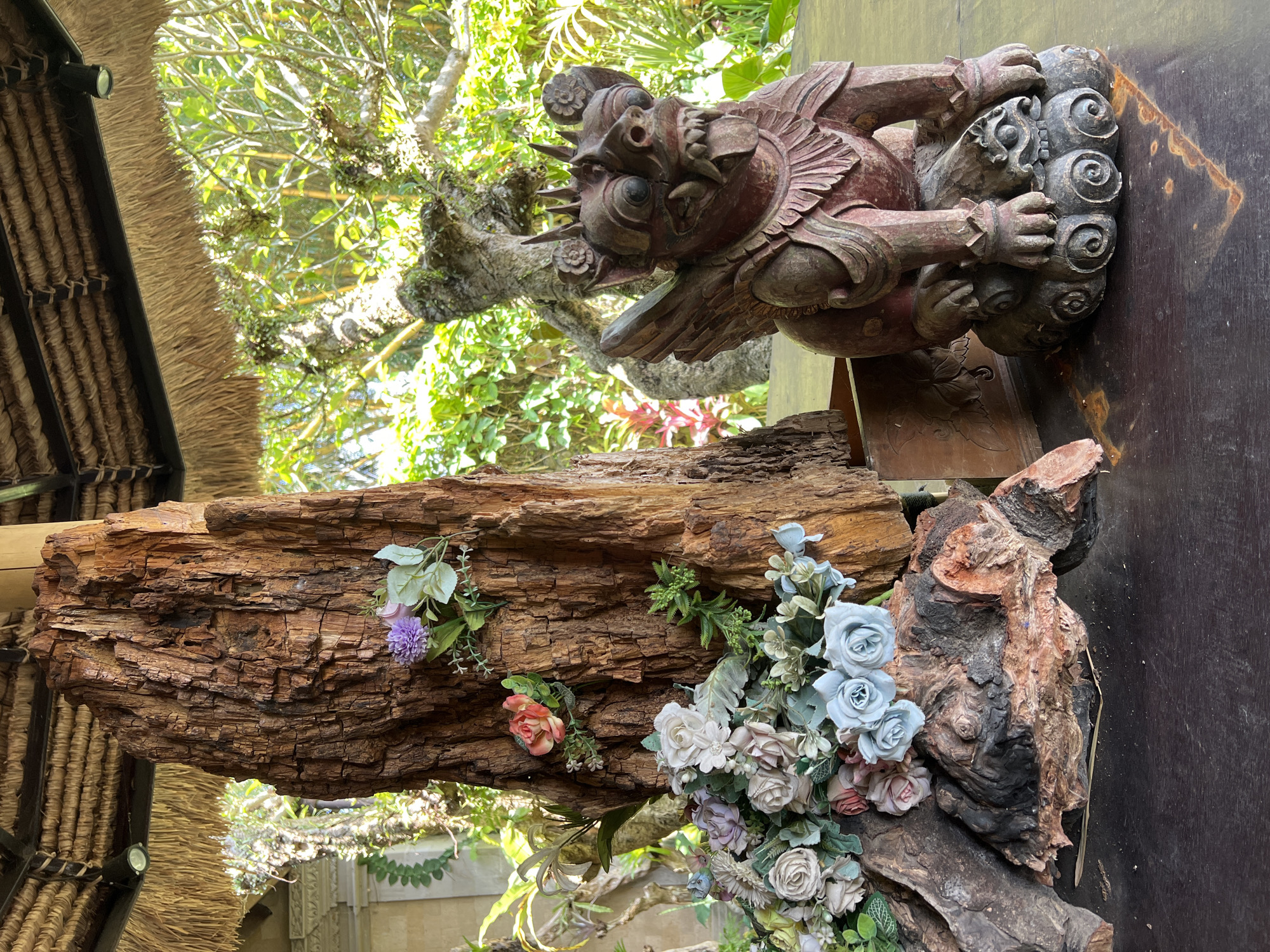 A wooden sculpture of a creature perches on a large piece of wood adorned with artificial flowers. The setting is likely outdoors, with elements of a thatched roof structure and lush green foliage visible in the background, aligning with the location of Kedewatan, Indonesia. The sculpture appears to be of a traditional style, possibly representing a guardian figure. The flowers are clustered primarily at the base of the wood and sparsely along its side, suggesting a decorative arrangement. The wood piece itself shows significant texture and cracks, indicating its age and exposure to the elements. The background features trees and some architectural details, pointing towards a garden or courtyard setting.