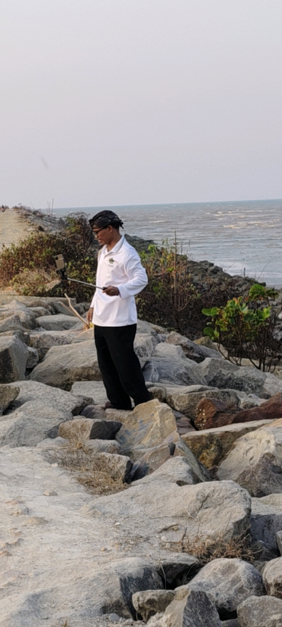 In Tasikagung, Indonesia, a man stands on a rocky shoreline, holding a selfie stick with a phone attached. He is wearing a white long-sleeved shirt and black pants. The sea is visible in the background, with small waves breaking. The sky is overcast. The man appears to be taking a photo or video of himself. The rocks are large and irregular, some covered with vegetation. A pathway is visible in the distance.