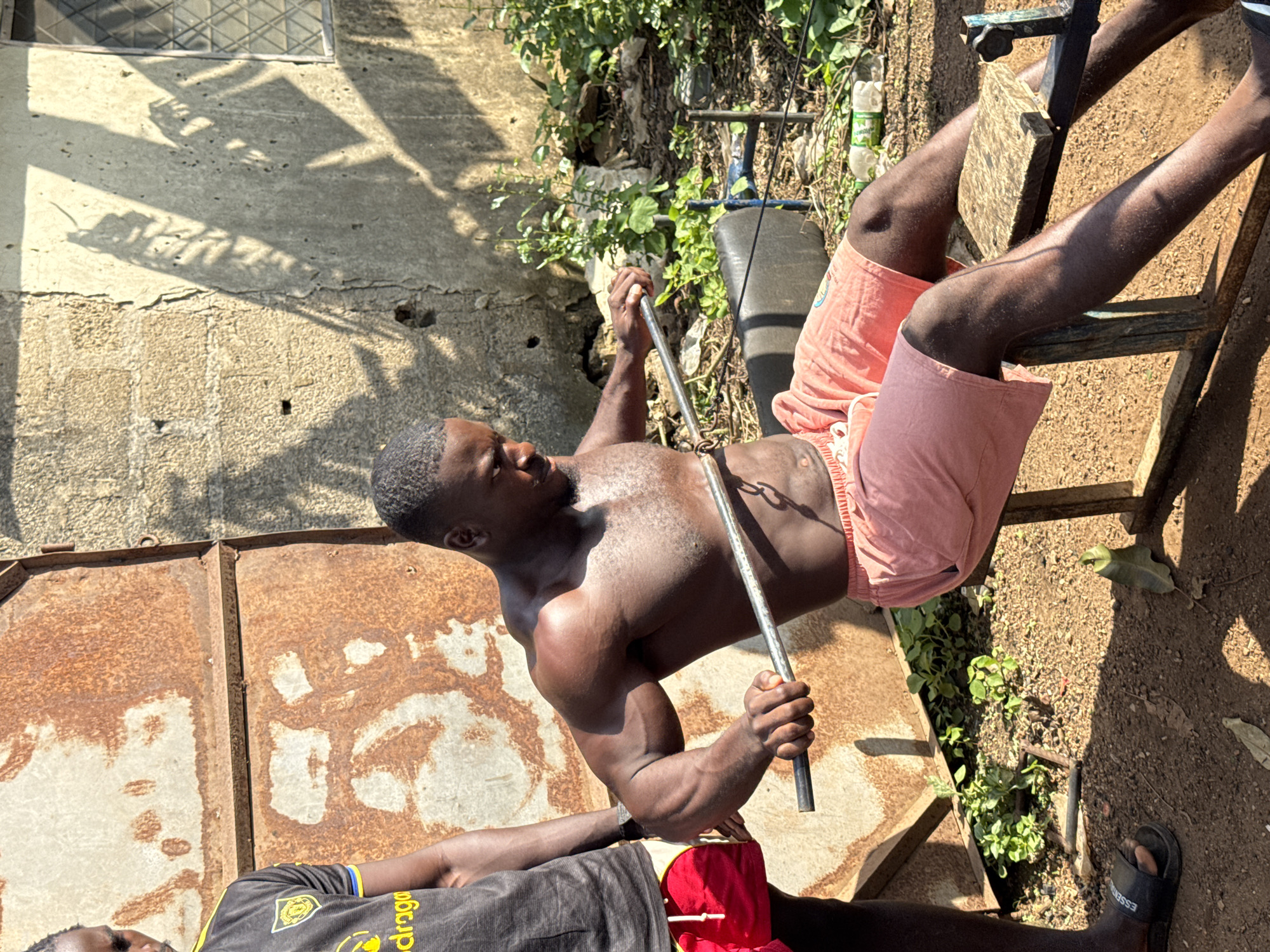 The media file depicts two individuals outdoors in what appears to be a rural or semi-urban setting in Akwanga, Nigeria. The primary subject is a muscular Black man performing an exercise, possibly a lat pulldown or a similar resistance exercise, using a metal bar attached to a weight system. He is shirtless, wearing light pink shorts, and his focus is intense. The scene is bathed in bright sunlight, suggesting it is daytime, likely mid-morning or afternoon, with distinct shadows cast on a concrete wall and rusted metal structures.
Behind the man exercising, there is a weight machine with a blue frame and a black padded bench. Green vegetation and some debris are visible in the background, indicating an outdoor area that is possibly a yard or an unkempt training space. To the left of the main subject, another person is partially visible, lying down and wearing a black t-shirt with yellow trim and writing on it. The t-shirt has a circular logo and the word "Dragona" is partially visible in yellow lettering.
The overall impression is one of dedication to physical fitness in a humble, resource-limited environment. The rusted metal surfaces and concrete wall suggest a potentially weathered or older structure, common in many Nigerian settings. There are no clear indicators of specific weather conditions beyond the bright sun, but the presence of foliage suggests a generally warm climate. The mood appears focused and determined, centered around the physical exertion of the main subject.