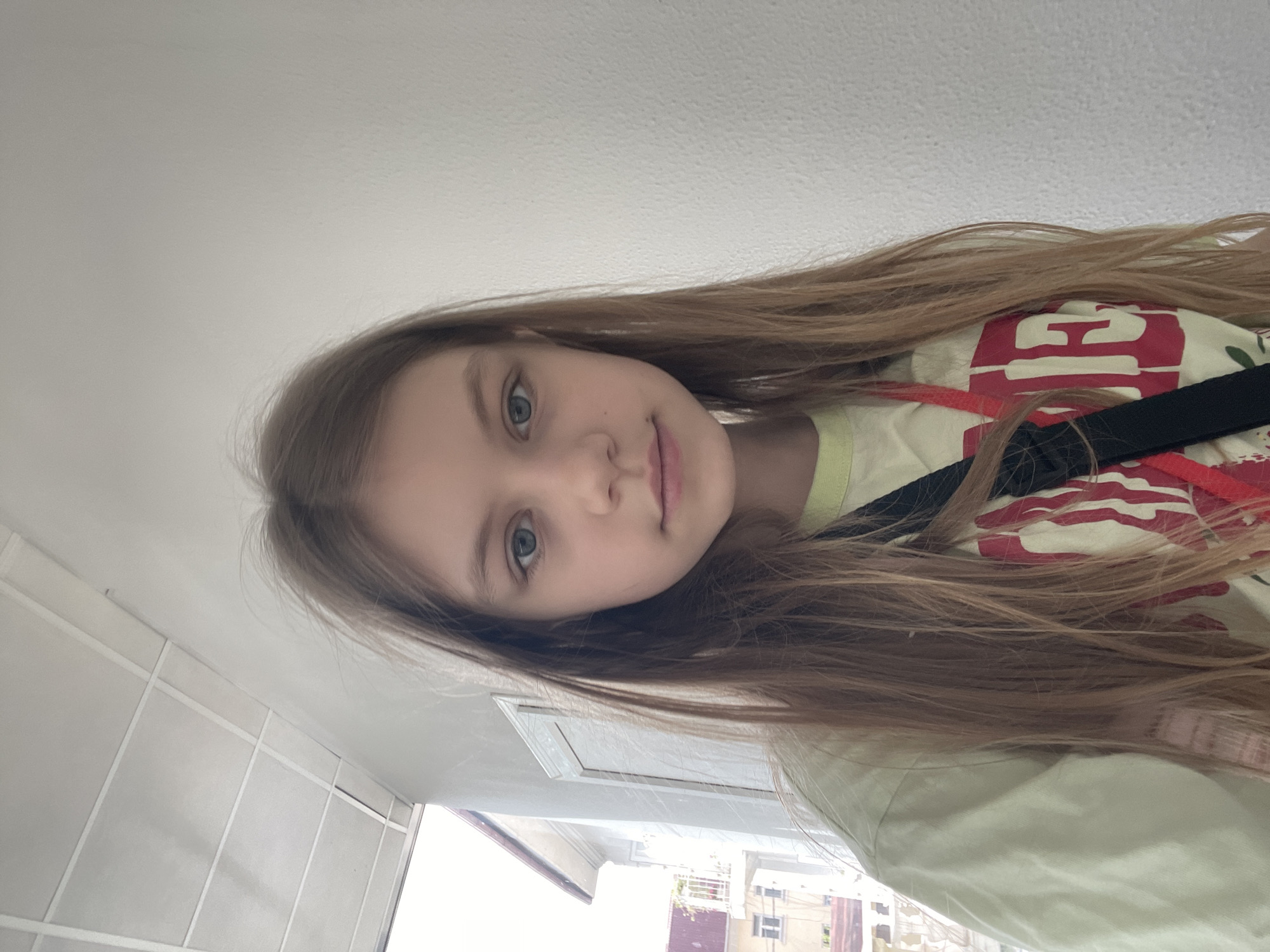A young girl with long, light brown hair and blue eyes is centered in the image, facing the camera. She has fair skin and is wearing a light-colored t-shirt with a red and white graphic on it, and a black strap is across her shoulder. The background includes a white ceiling and a tiled wall to the left. Visible through a window is a cityscape, including buildings with light-colored facades and red roofs. The location is Sochi, Russia.