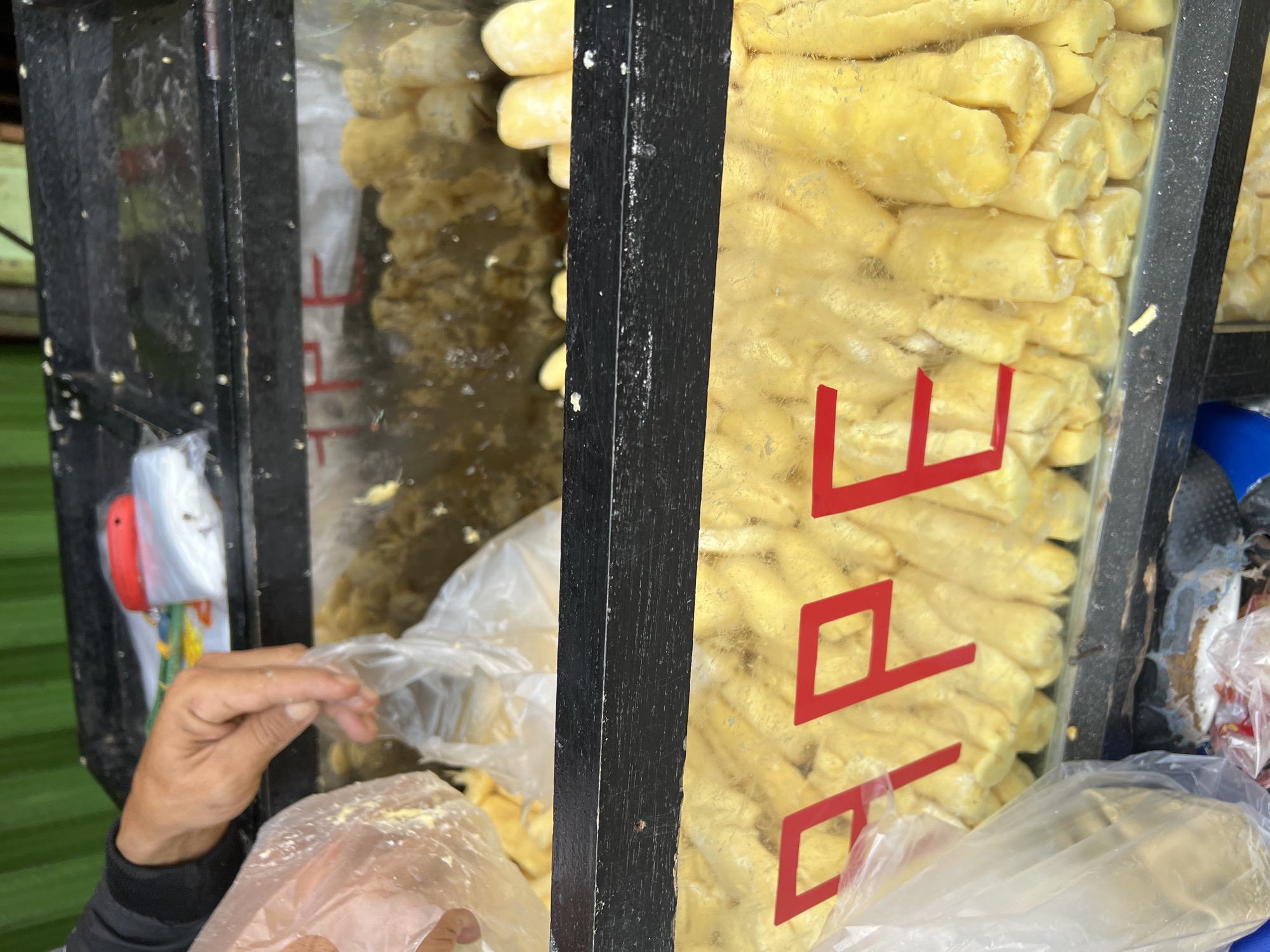 In Citeureup, Indonesia, a close-up, eye-level shot captures a vendor's hands reaching into a display case filled with yellow food items. The display case is divided into two visible compartments by a black horizontal bar. The upper compartment contains a blurred assortment of food items and plastic bags, with a red object and what appears to be a knife handle visible on the top surface. The lower compartment is more distinctly filled with numerous yellow, cylindrical food items, possibly fried snacks or pastries, arranged vertically. Red lettering is adhered to the glass of the lower compartment, partially obscuring the food; the visible letters spell "APE". A person's hands, one holding a clear plastic bag and the other manipulating another plastic bag, are in the process of handling food. The background is blurred and shows a green corrugated surface, likely a wall or awning. The overall scene suggests a street food vendor or a market stall setting.