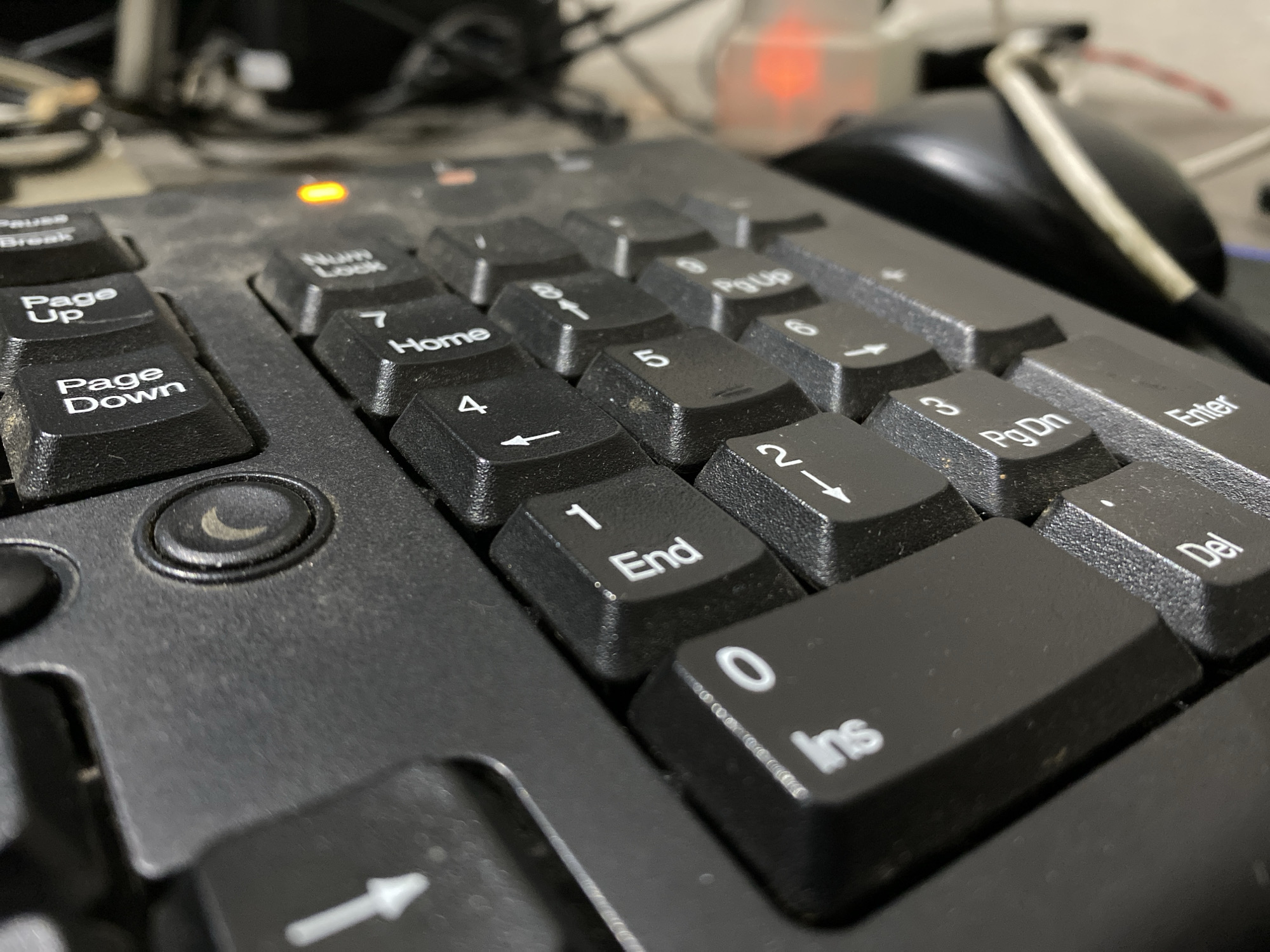 The image is a close-up, low-angle shot of a dusty black computer keyboard. The focus is on the numeric keypad section. Visible keys include "Pause Break," "Page Up," "Page Down," "Num Lock," "7 Home," "4," "1 End," "0 Ins," "Pg Dn," and "Enter." There is an orange LED light illuminated on the keyboard, likely indicating "Num Lock" is active. The background is blurred and shows electronic equipment and cables. The scene is indoors, suggesting a workspace. The image was captured in Dabasah, Indonesia. There are no people or discernible actions taking place.
