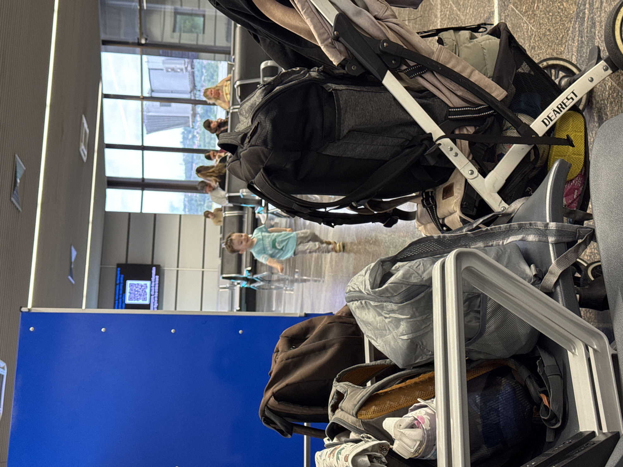 The image shows an indoor scene, likely an airport waiting area. In the foreground, there's luggage on a luggage cart and a stroller, obscuring much of the view. People are seated in the background near a large window with a cityscape visible through it. A child is running in the middle ground. A QR code display is seen in the background. The stroller's brand is visible and reads "DEAREST". I cannot confirm the location as Sochi, Russia.