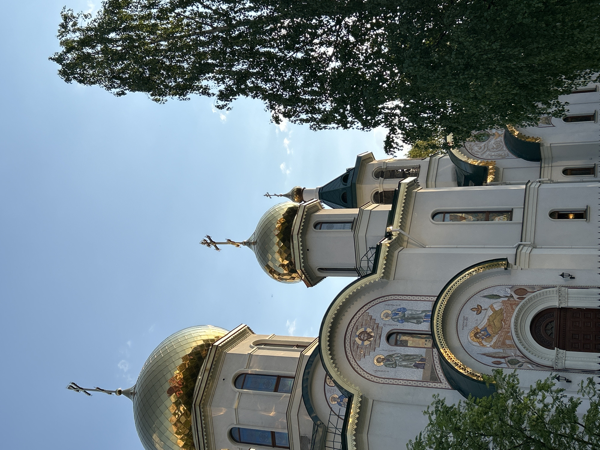 This image captures a low-angle view of the Transfiguration Cathedral, located in Saint Petersburg, Russia. The cathedral's architecture features prominent golden domes topped with Orthodox crosses, juxtaposed against a clear blue sky and the foliage of a tree overhead. The building's exterior is adorned with religious mosaics and decorative elements, indicative of traditional Russian Orthodox design. The scene is brightly lit, suggesting daytime conditions.