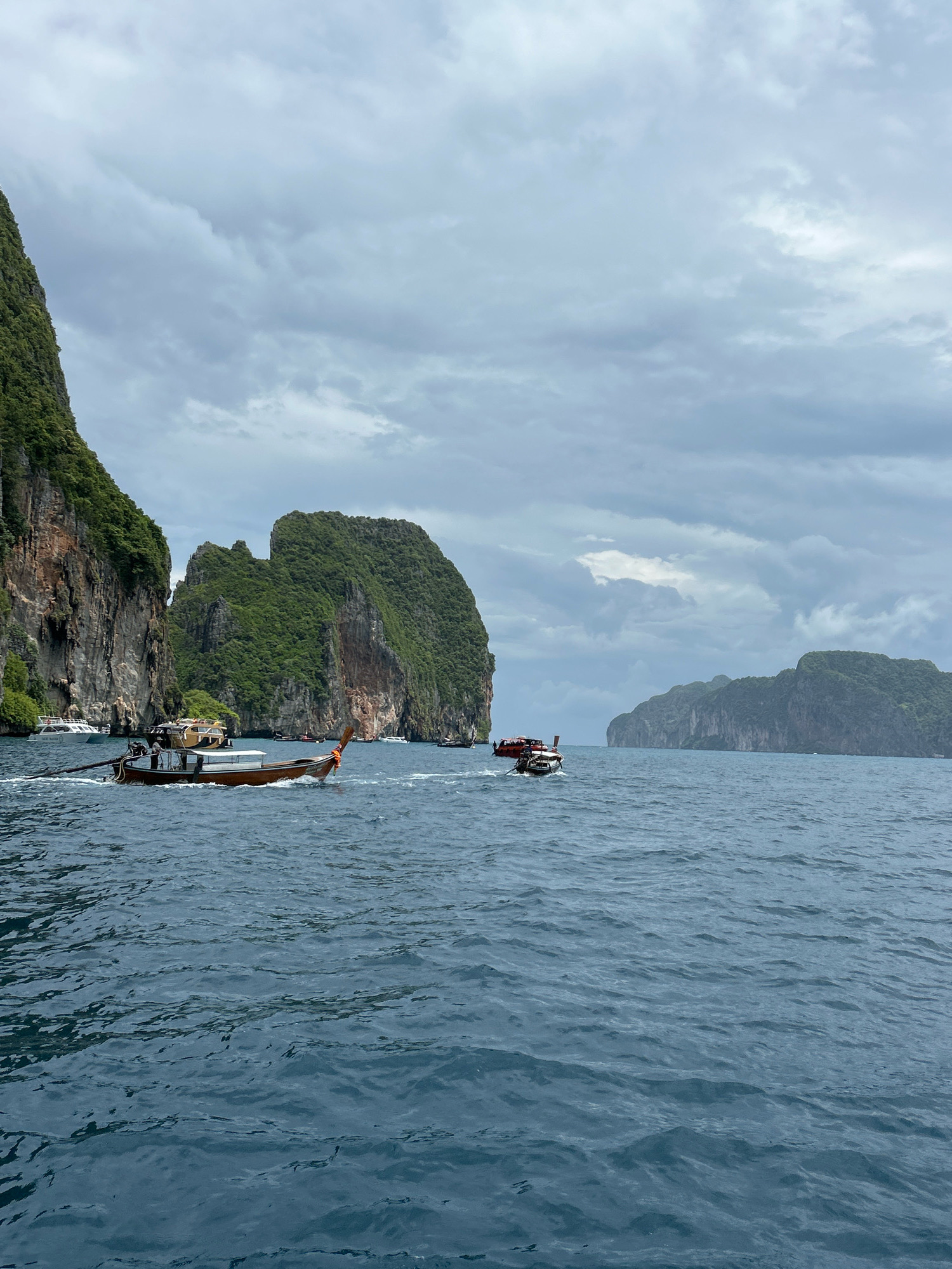 This image captures a scenic view of the Andaman Sea near Ao Nang, Thailand, under a cloudy sky. In the foreground, a traditional longtail boat navigates the choppy, dark blue water. Further out, another longtail boat with a red hull is visible, as are several other boats of varying sizes, some appearing to be speedboats. The background is dominated by dramatic limestone karst islands, characterized by their steep cliffs covered in lush green vegetation. These islands rise majestically from the sea, creating a breathtaking natural landscape. The overall atmosphere is one of natural beauty and marine activity, typical of the Ao Nang region.