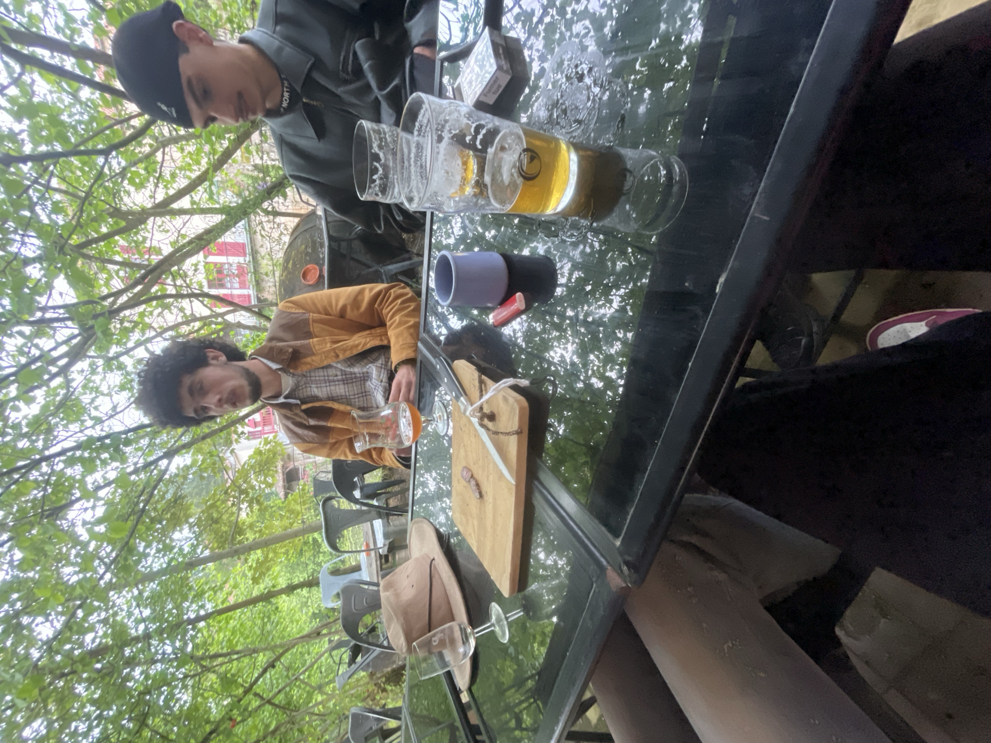 Two individuals sit at a black-topped table outdoors, presumably at a café in Parthenay, France. A younger male wearing a black cap and gray jacket is seated at the head of the table, while another male with curly dark hair and a brown jacket sits perpendicular to him. On the table, there is a beer glass, a mug, a cutting board with a knife, a hat, and two wine glasses. The scene is set in a green, wooded area with visible buildings, suggesting the location is in the city of Parthenay.