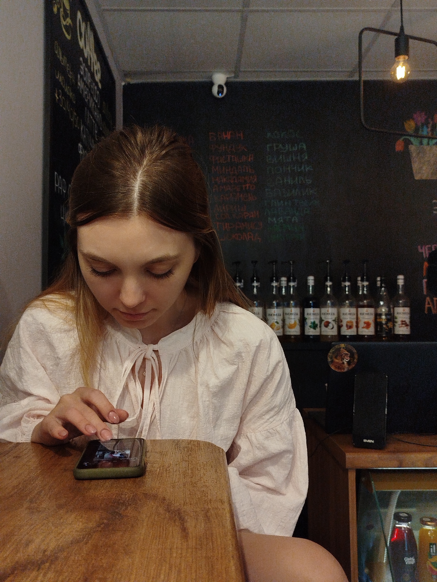 A young woman with long brown hair, wearing a light pink blouse with a tie-neck, sits at a wooden table in what appears to be a coffee shop. She is looking down at her smartphone, which she is holding in her left hand. Her right hand is poised over the screen, suggesting she is interacting with it. The background features a chalkboard with handwritten text and illustrations, a security camera, a hanging light fixture, a row of syrup bottles, a black speaker, and a shelf with bottles of beverages. The overall environment suggests a casual, indoor setting.