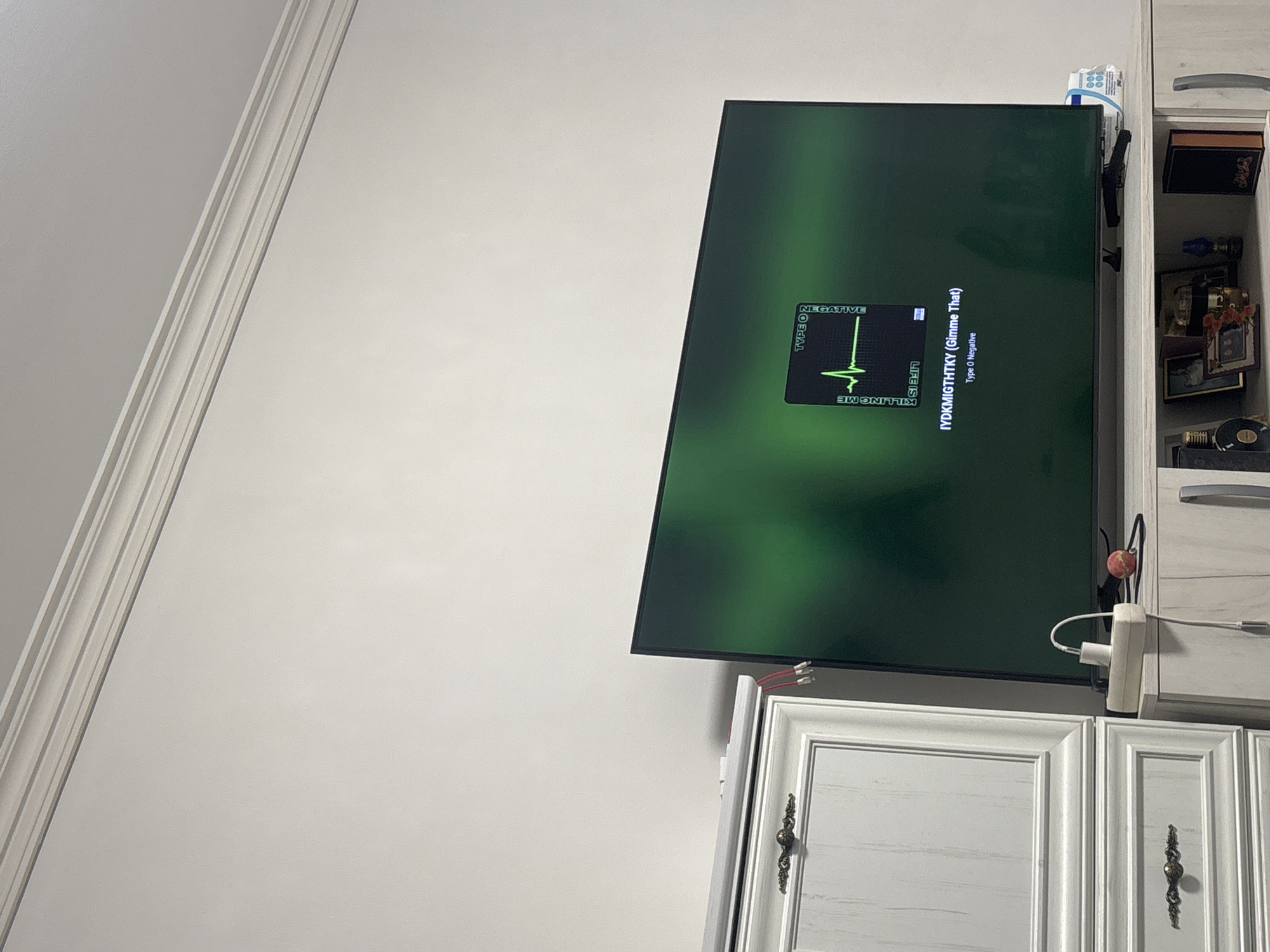 The image depicts a close-up of a television screen displaying album art for the song "IYDGMIGTHTKY (Gimme That)" by Type O Negative. The album art features a stylized electrocardiogram on a black background with green text. The television is positioned on top of a light-colored wooden entertainment center. To the left of the television is a tall, light-colored cabinet with ornate brass drawer pulls. Several wires and cables are visible connected to the television and the entertainment center. A white power strip with a white plug is also on the entertainment center, along with a small red apple and a black object resembling a handgun. The background consists of a plain, light-colored wall with decorative molding at the top. The scene appears to be an indoor setting, possibly a living room or entertainment space. The image is taken from a slightly low angle, looking up towards the television and the wall.
