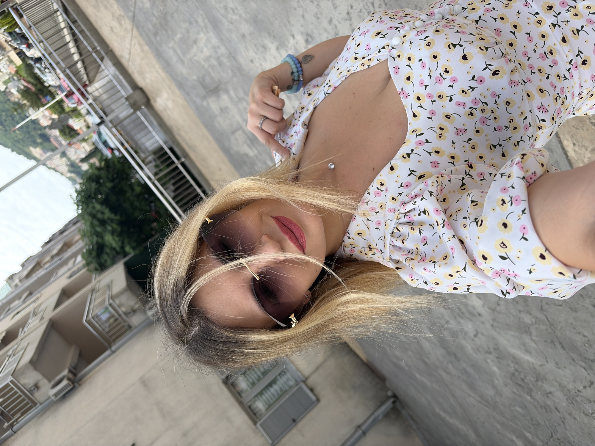 This media file presents a close-up, tilted selfie of a woman in an outdoor setting. The woman has blonde hair with visible darker roots and is wearing stylish sunglasses with dark lenses and golden accents. Her lips are adorned with pink lipstick, and she has fair skin. She is dressed in a white short-sleeved dress featuring a cheerful pattern of small yellow and pink flowers with black details, and a button-up front. Her accessories include multiple light blue and turquoise beaded bracelets on her right wrist, a ring on her left ring finger, and a subtle dermal piercing with a clear jewel on her upper chest/collarbone area. A small leaf-like tattoo is visible on her right forearm.
The background reveals an urban environment, likely a coastal town such as Budva, Montenegro. Multi-story buildings with numerous balconies and windows are visible, suggesting residential or hotel structures. In the upper left corner, beyond some metallic railings or staircase structures, a wider vista of distant buildings and natural landscape or possibly a body of water can be discerned. Trees and foliage are scattered throughout the background. The lighting suggests it is daytime, with bright, even illumination and clear weather. The woman appears relaxed, looking towards the camera as she takes the photo. There is no visible text in the image.