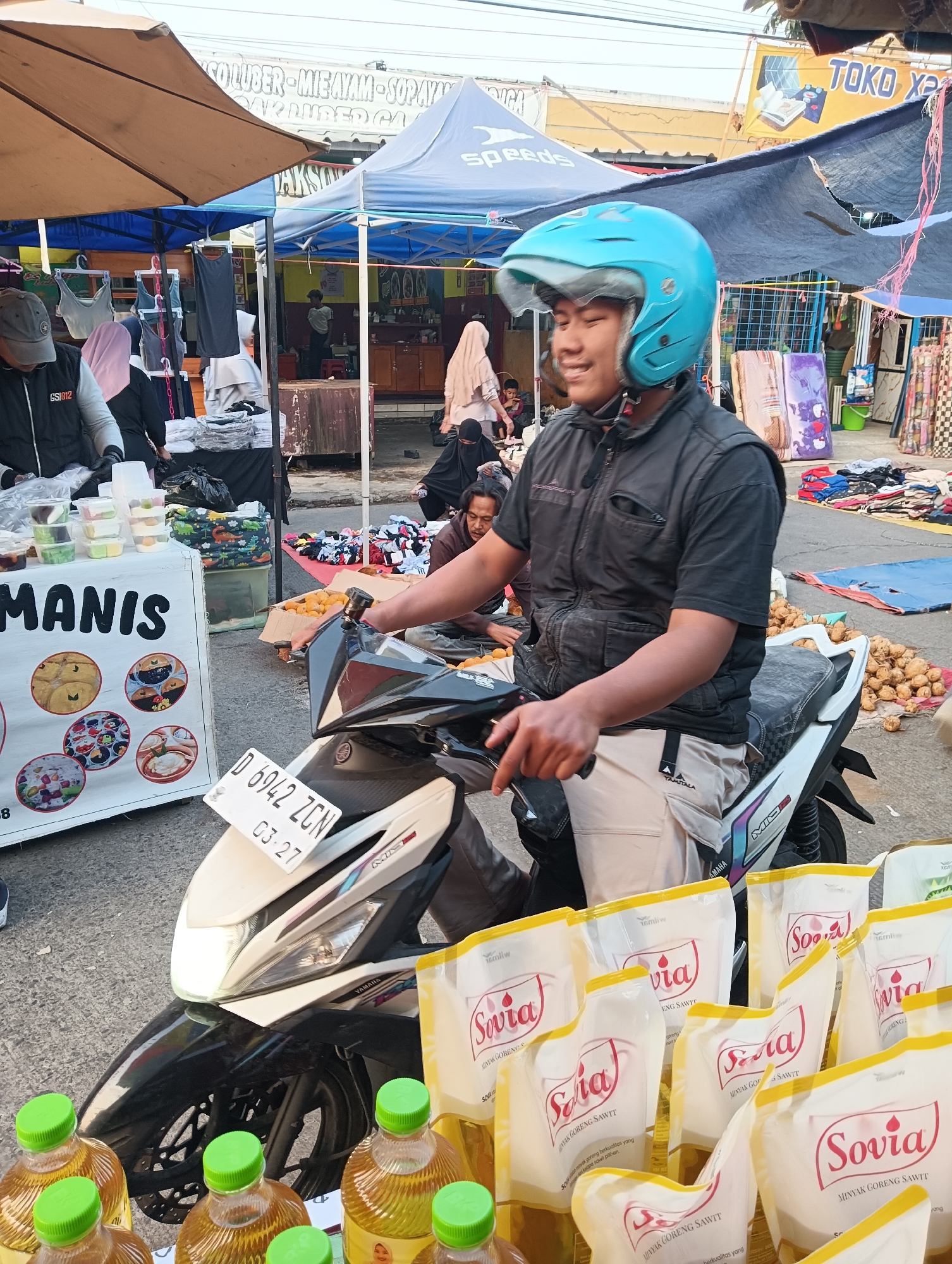 In the city of Ketapang, Indonesia, a street market scene unfolds under an overcast sky. A young man, wearing a bright blue helmet and a dark vest over a light-colored shirt and pants, sits astride a white and black motorcycle. His right hand rests on the handlebar, and he gazes to his right with a slight smile. The motorcycle's license plate reads "D 6942 ZCN 03-27".
In the foreground, several bottles with green caps are partially visible, their contents appearing to be a yellowish liquid. Behind these bottles are multiple pouches, some displaying the "Sovia" brand name and "Minyak Goreng Sawit" (Palm Cooking Oil).
The market itself features various stalls. To the left, a white stand with colorful circular images advertises food items, with the word "MANIS" (sweet) prominently displayed. Further back, various items are displayed under blue and tan awnings, including clothing and what appear to be pillows or cushions. Several people are visible in the background, some tending to stalls or browsing goods. The ground is paved, suggesting a street or walkway within the market area.