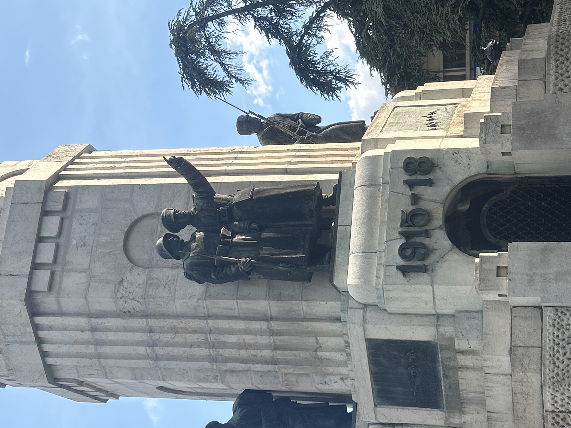 The image shows a war memorial in Veliko Tarnovo, Bulgaria. The monument is made of light-colored stone and features bronze sculptures of soldiers in different poses. The years "1915-1918" are inscribed on the side of the memorial. The sculptures depict soldiers advancing, one pointing forward, and another lying down with a rifle. There are trees and a blue sky in the background.