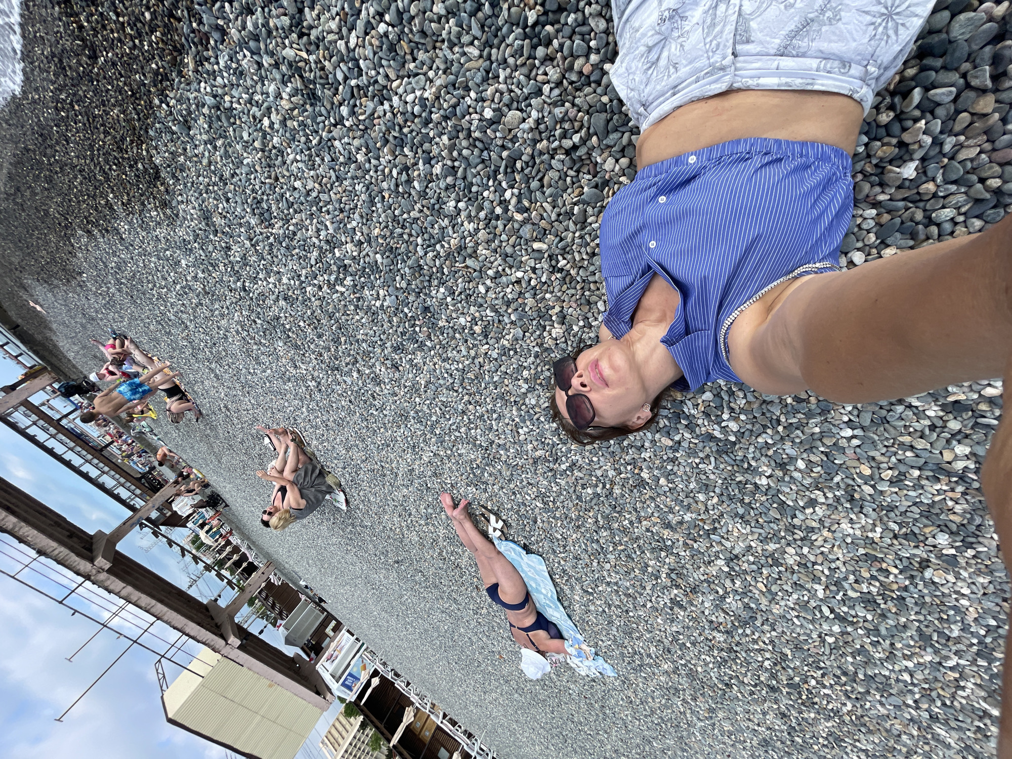 A selfie taken from a low angle on a pebble beach shows a woman with sunglasses and a blue and white striped shirt lying down, with her arm extended. She is wearing patterned shorts. To her left, a person in a blue bikini and white hat lies on a blue towel, facing away from the camera. In the background, other people are visible on the beach, some sitting and some standing. A large, concrete structure, possibly a pier or bridge, runs overhead. Buildings and trees can be seen in the distance. The water is visible at the top of the image. The scene appears to be a beach resort area in Sochi, Russia.