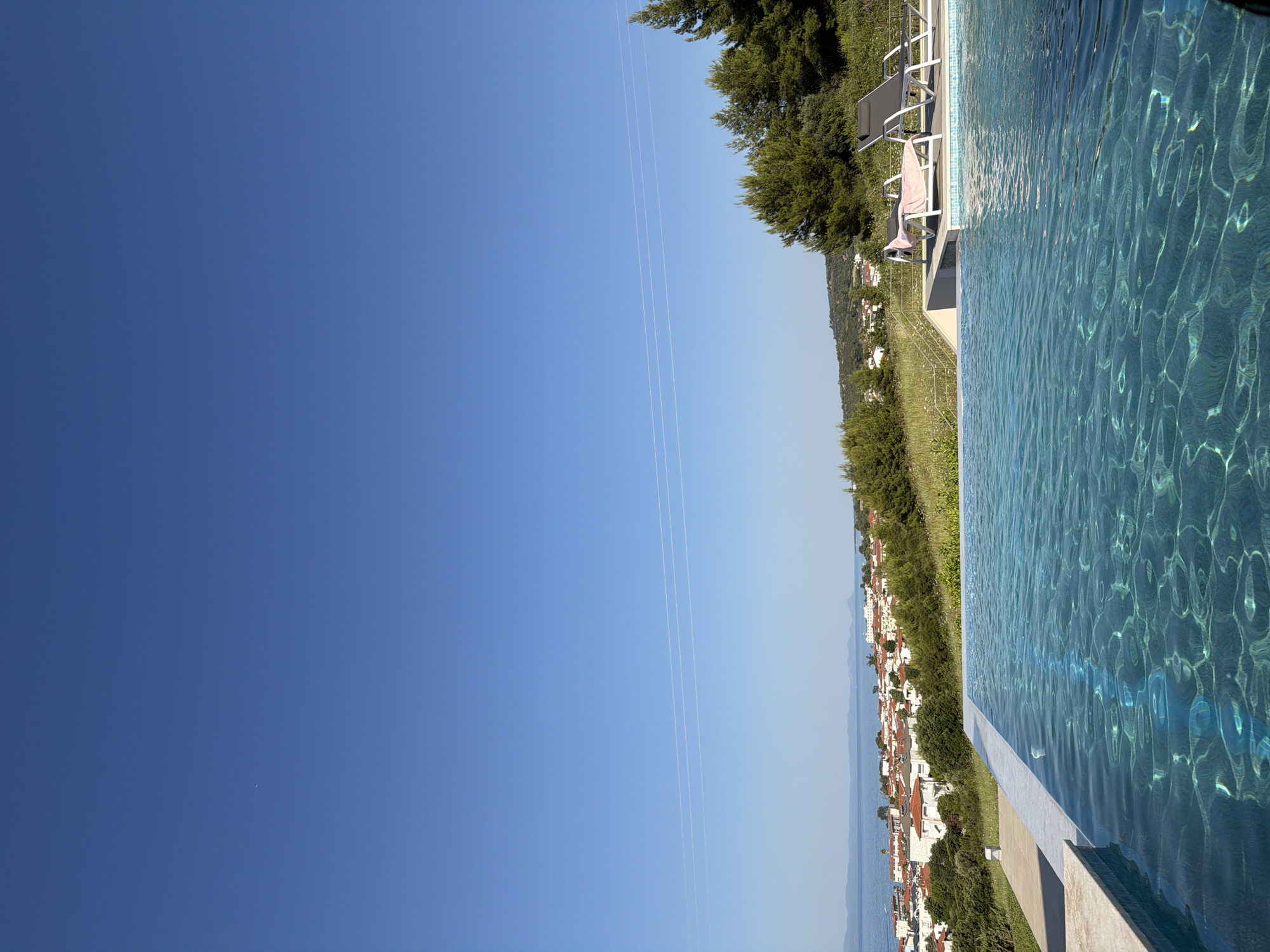 This is a landscape photograph of a tranquil scene in Pallini Municipal Unit, Greece. The image features a clear blue sky dominating the left side, with three faint contrails stretching across it. To the right, a pristine swimming pool with rippling, turquoise water takes up a significant portion of the frame. The pool's edge is visible, with a light-colored coping and a greyish concrete base.
Beyond the pool, a hillside slopes down towards a coastal town. The town is characterized by clusters of white buildings with red-tiled roofs, nestled amidst lush green vegetation. Trees are abundant on the slopes, and further in the distance, the calm sea is visible, merging with the hazy horizon.
On the deck of the swimming pool, two white lounge chairs are placed, one with a pink towel draped over it, suggesting a moment of relaxation and leisure. The scene evokes a sense of peace and summer vacation. The bright, clear sky indicates it is daytime, likely a sunny afternoon. No people are explicitly visible in the frame. There is no visible text in the image.