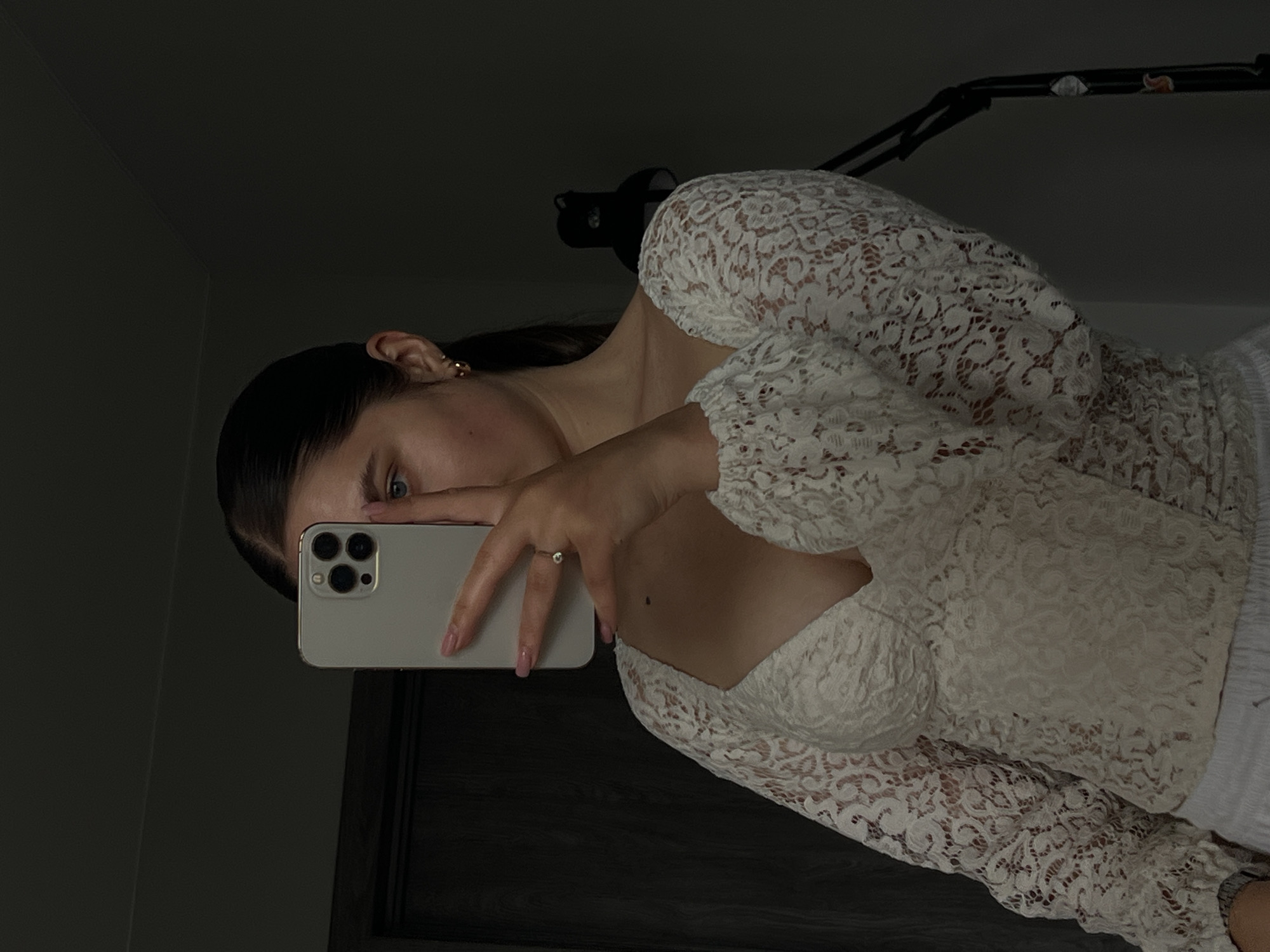 The image displays a young woman taking a mirror selfie in a dimly lit indoor environment, likely located in Ternopil, Ukraine. She holds a light-colored smartphone horizontally in front of her face, partially obscuring it, with only her left eye and forehead visible. Her gaze is directed towards the phone, suggesting she is focused on taking the picture.
The woman has dark hair, which appears to be pulled back. She is wearing an elegant white lace top with long sleeves and a sweetheart neckline, revealing a hint of décolletage. A delicate ring with a single small stone adorns her left ring finger, and her fingernails are painted with light pink polish. Small, gold-colored hoop earrings are visible on her left ear.
The background is dark and largely indistinct, with glimpses of what might be a dark door frame or wall in the lower right, and a multi-jointed dark object, possibly a lamp arm or tripod, extending across the upper part of the frame. The low light levels give the image a moody, intimate feel, with the primary illumination appearing to come from an unseen source, possibly the phone screen itself or a soft ambient light. There is no visible text in the image. The scene captures a personal, casual moment.