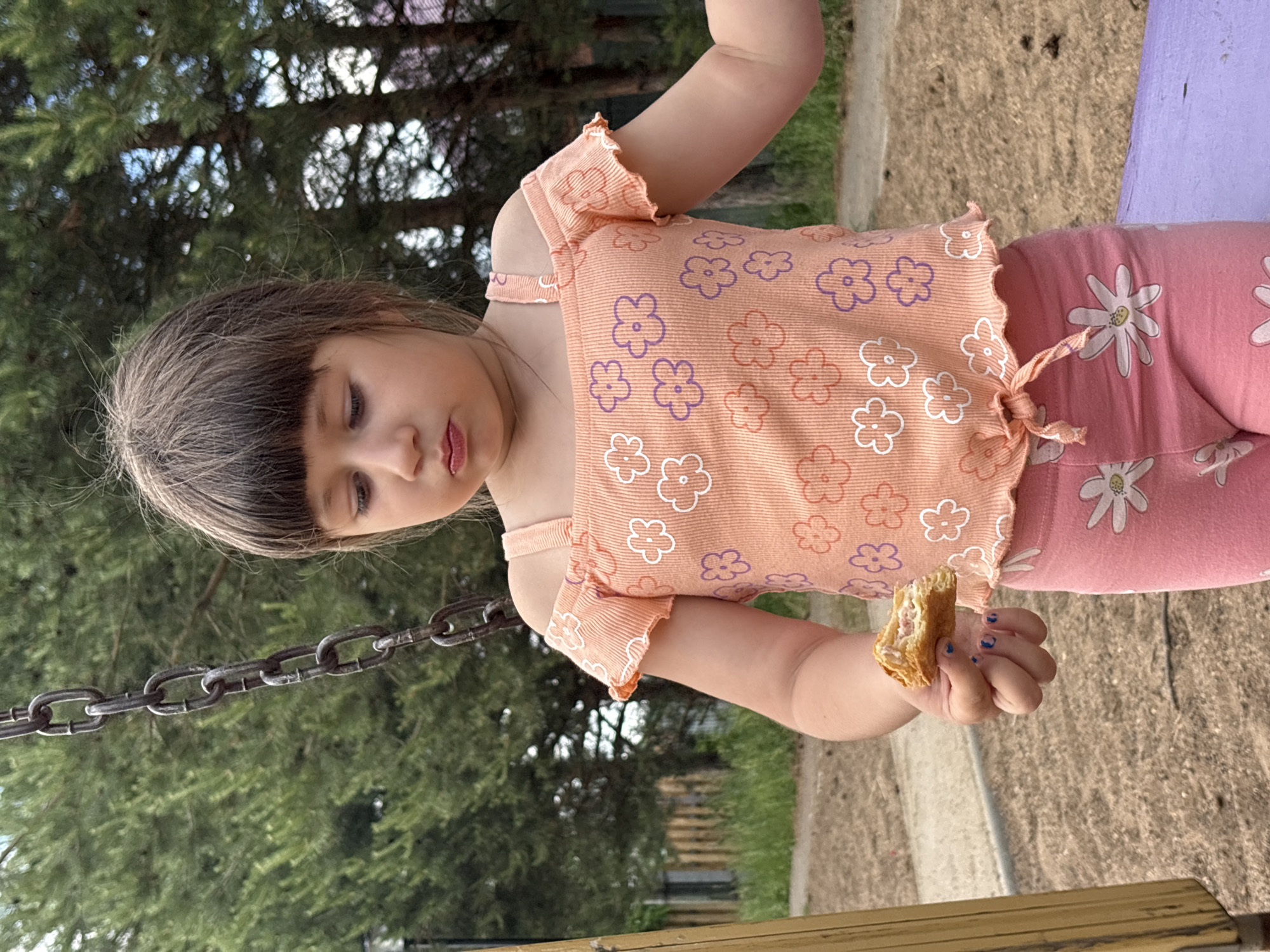 A young girl with brown hair and bangs is standing outdoors, holding a piece of food in her right hand. She is wearing an orange, off-the-shoulder top with a floral pattern and pink leggings adorned with white flowers. The girl is looking down at the food she is holding. Behind her, a metal chain hangs from above, and trees and a wooden fence are visible in the background. The ground appears to be covered in sand or dirt. The scene is likely at a playground, given the presence of the chain and the overall environment. This image was captured in the city of Chaikovsky, Russia.