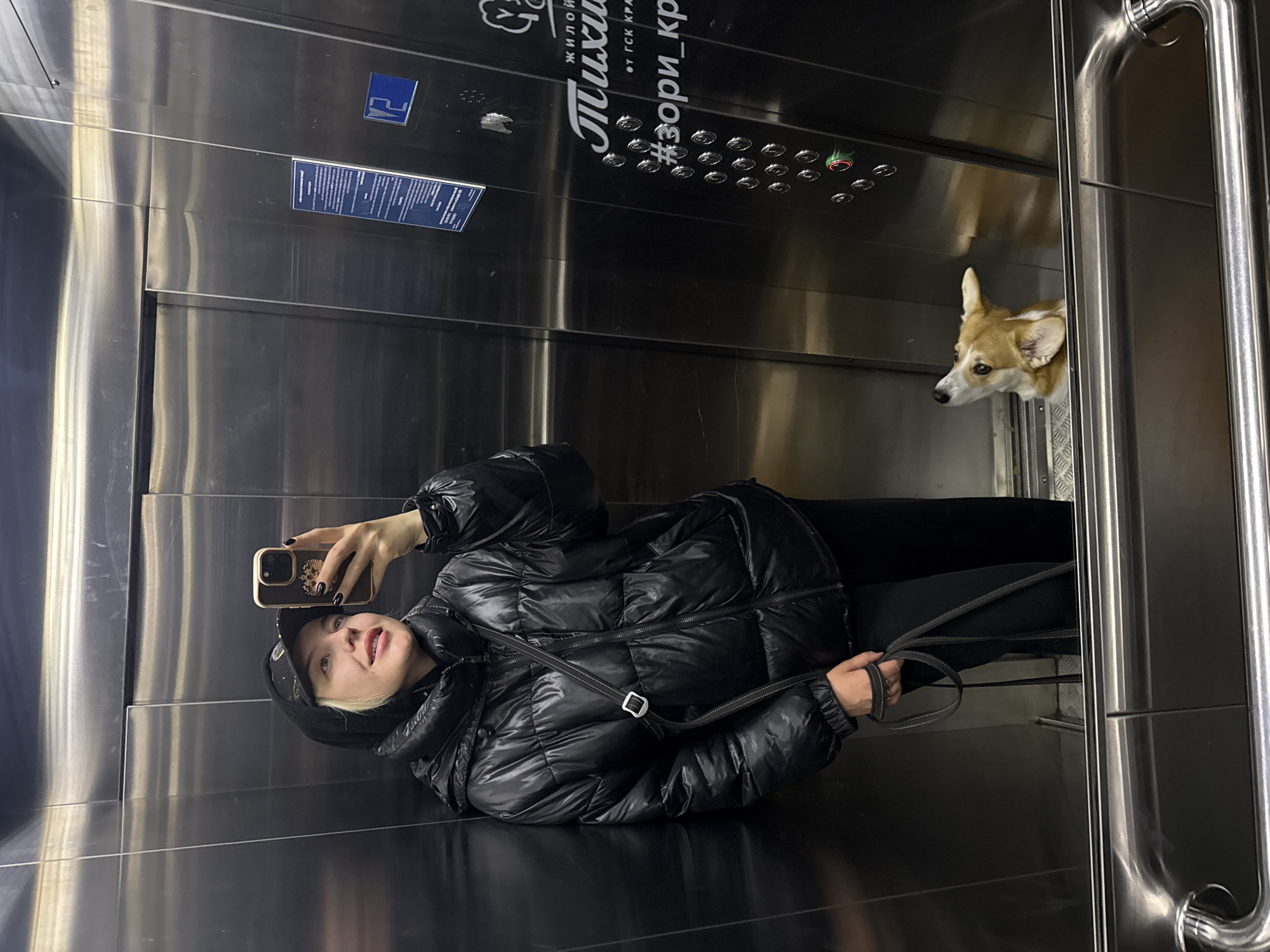 A woman is taking a selfie in the mirror inside an elevator. The elevator is located in Krasnoyarsk, Russia, as indicated by Cyrillic text on the elevator panel. The woman is wearing a black hooded puffer jacket and a hat. She is holding a phone with her right hand. A dog's head is visible in the reflection, looking out from the elevator car behind the woman. The elevator has stainless steel interior.