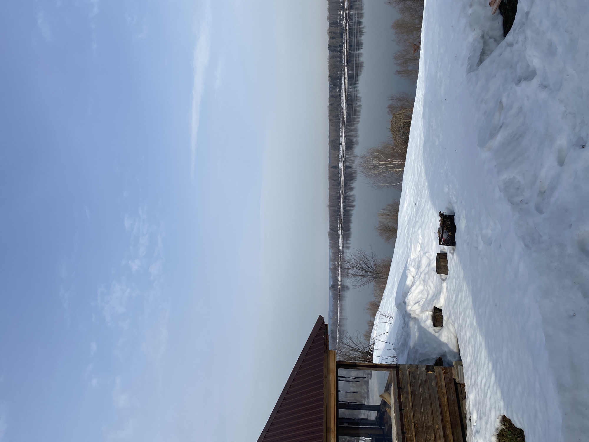 The image displays a winter or early spring landscape located in Ольховка, Russia. The scene is dominated by a wide, calm river occupying the middle ground, reflecting the bare trees of the distant riverbank. The sky above is clear light blue with minimal, faint wispy clouds. In the foreground, on the left, a rustic wooden pavilion with a dark reddish-brown corrugated metal roof is partially visible, supported by stone blocks and surrounded by snowdrifts. The ground between the pavilion and the river is covered in thick white snow, interspersed with patches of exposed brown earth and sparse, dormant vegetation. Two dark tree stumps and a dark, rectangular brazier are positioned on the snow-covered terrain near the pavilion. The river's surface appears dark and still, bordered by a dense line of bare deciduous trees on the opposite shore. No people are present in the scene.